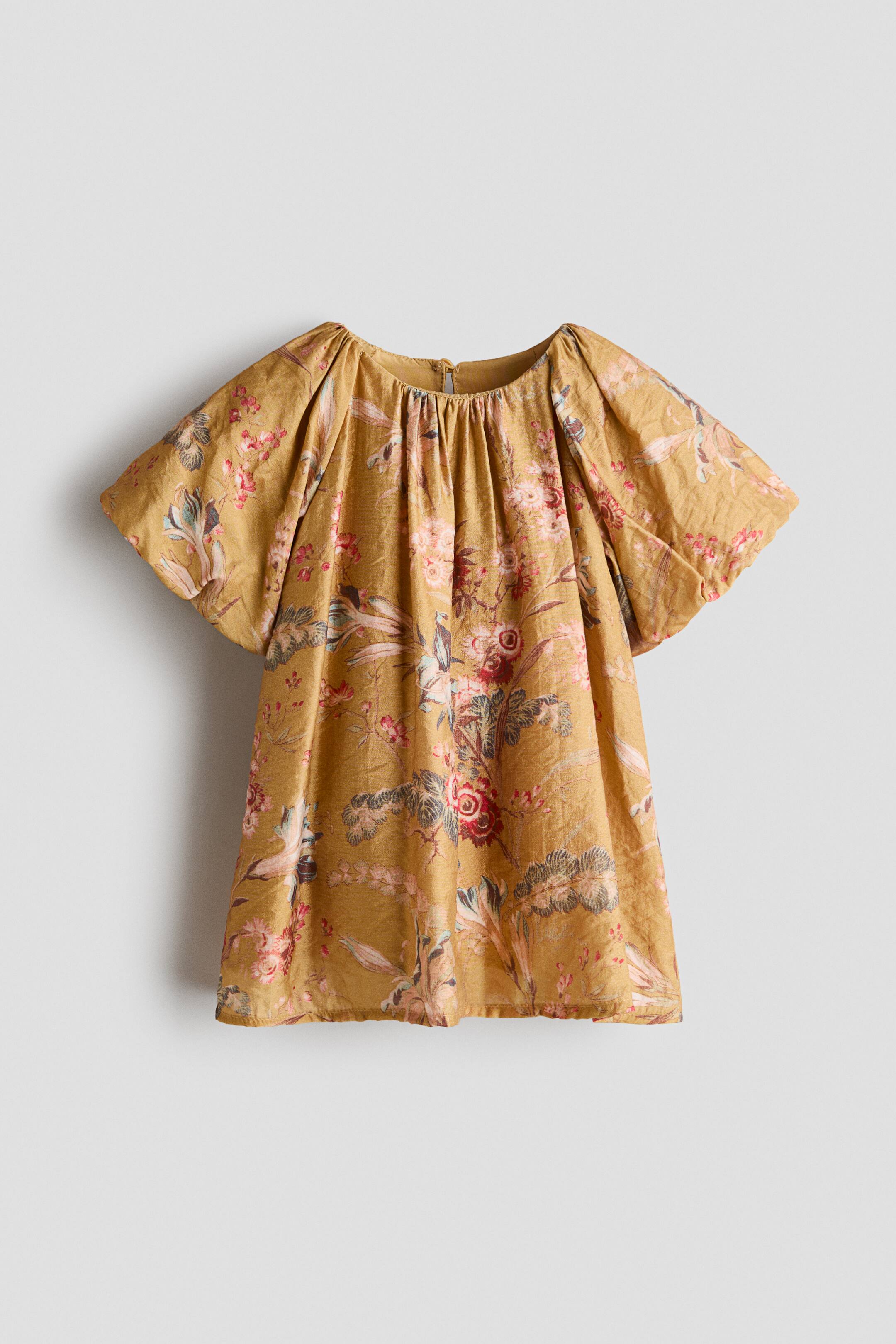 View larger image: A mustard-yellow blouse showcases an all-over vintage floral pattern in muted red, pink, and green tones. The top features a gathered round neckline, short, voluminous puff sleeves, and a loose, flared silhouette in a front-facing view.