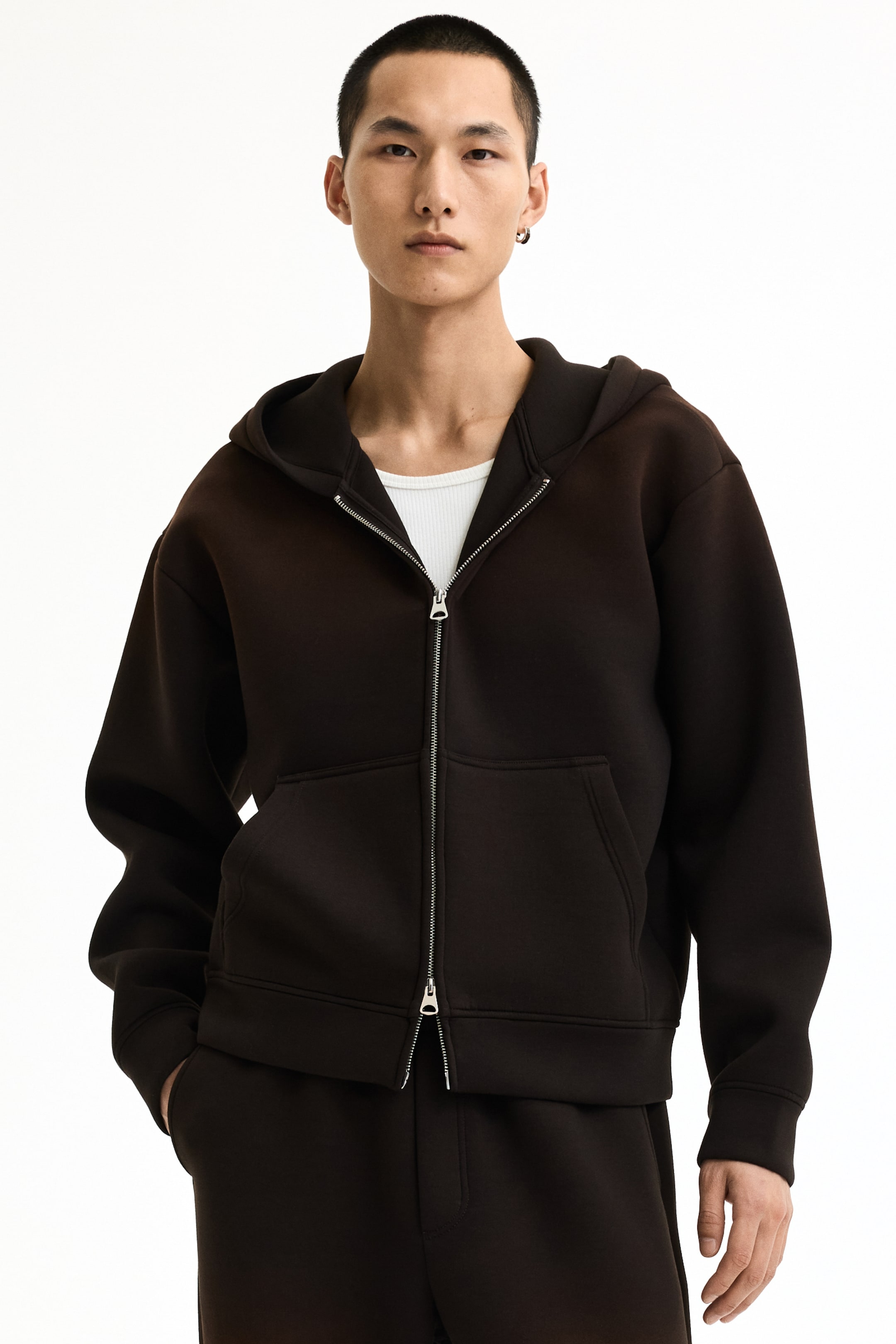 View larger image: A dark brown full-zip hoodie showcases a structured design, facing forward. It features a silver-toned full-length zipper, front kangaroo pockets, and a structured hood, worn partially open over a white ribbed tank top.