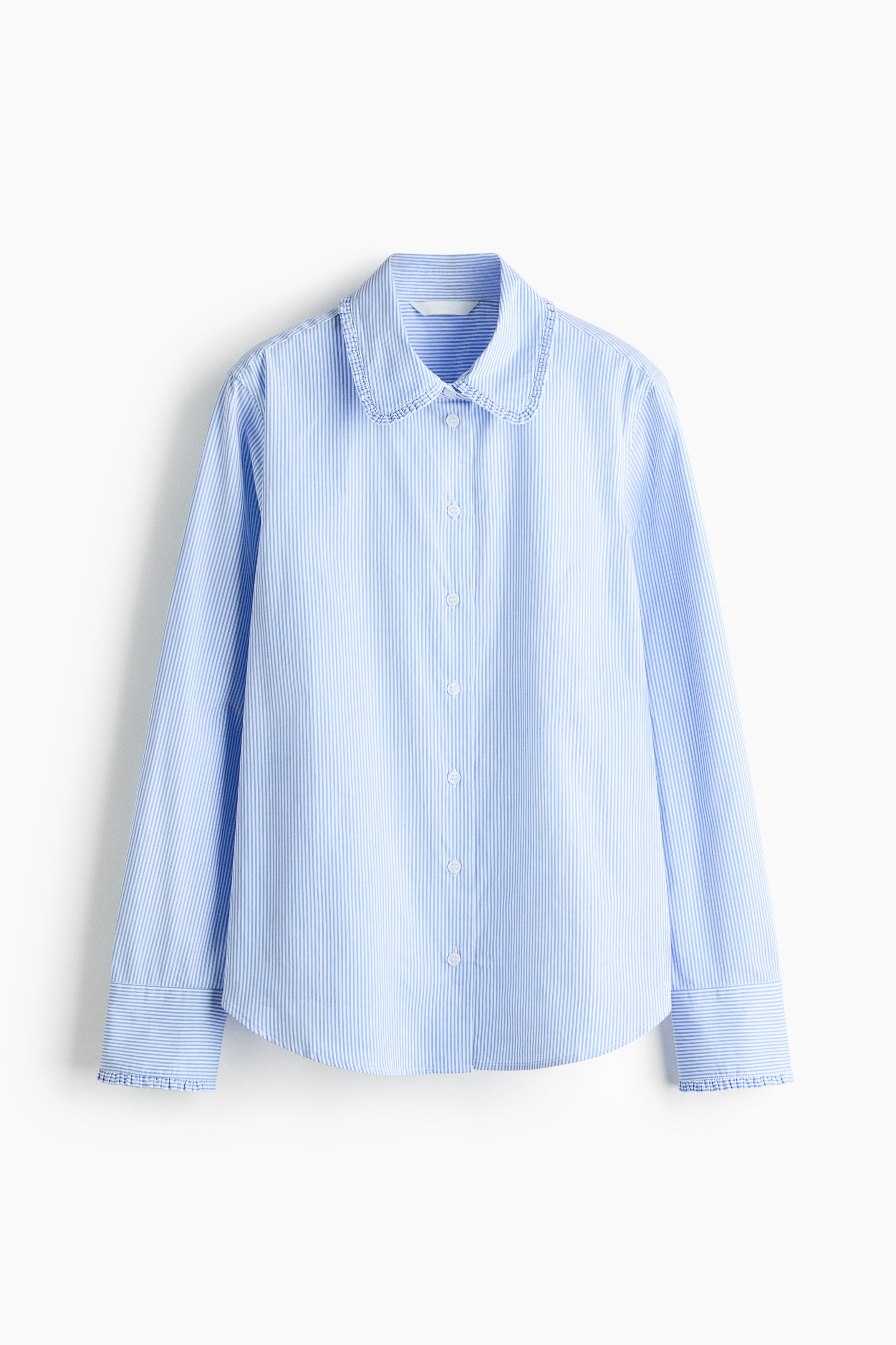A blue and white striped long-sleeve shirt featuring a frill-trimmed collar and cuffs is being worn open by the model, who is crouching. The shirt is layered over a white top and paired with white trousers.