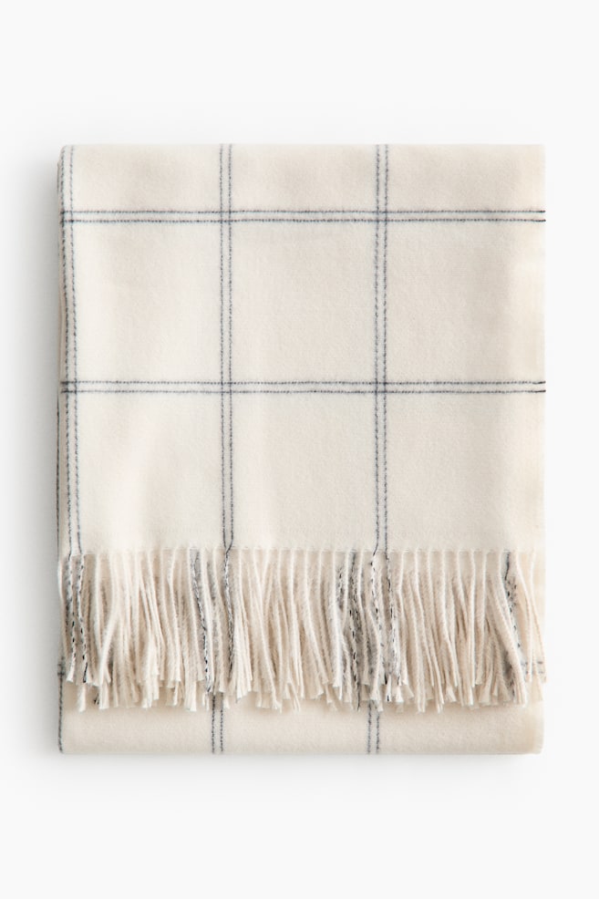 A cream blanket is neatly folded flat, showcasing a dark windowpane check pattern and featuring a fringed edge along its bottom. - 1