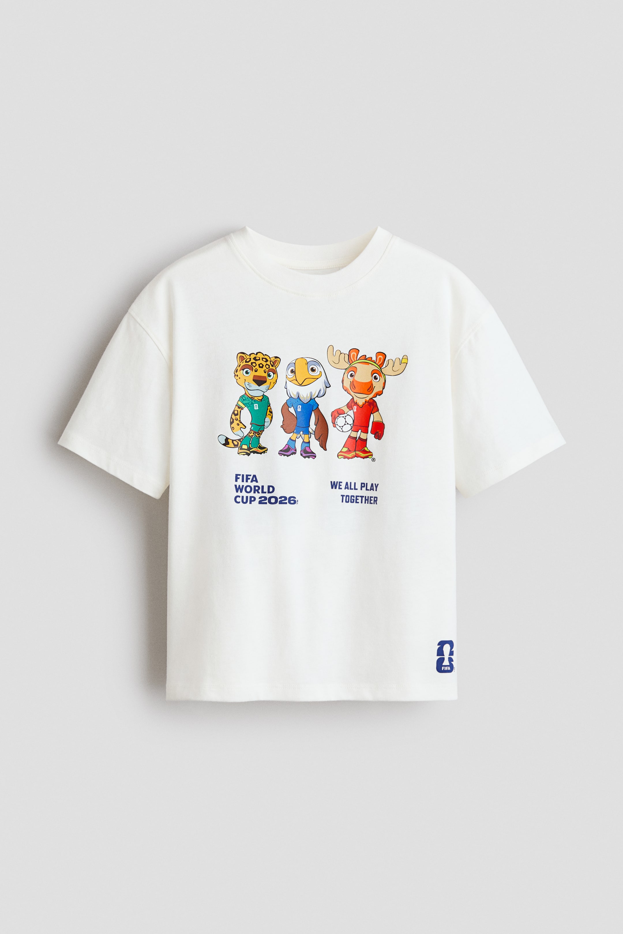 Größeres Bild anzeigen: Ein übergroßes, cremeweißes Baumwolljersey-T-Shirt zeigt auf der Vorderseite einen bunten Grafikdruck mit drei Cartoon-Maskottchen – einem Leoparden, einem Adler und einem Elch – in Fußballuniformen. Der Text lautet "FIFA WORLD CUP 2026" und "WE ALL PLAY TOGETHER". Es verfügt außerdem über einen gerippten Besatz am Rundhalsausschnitt, tief angesetzte Schultern, und ein kleines blaues FIFA-Logo erscheint am unteren rechten Saum.