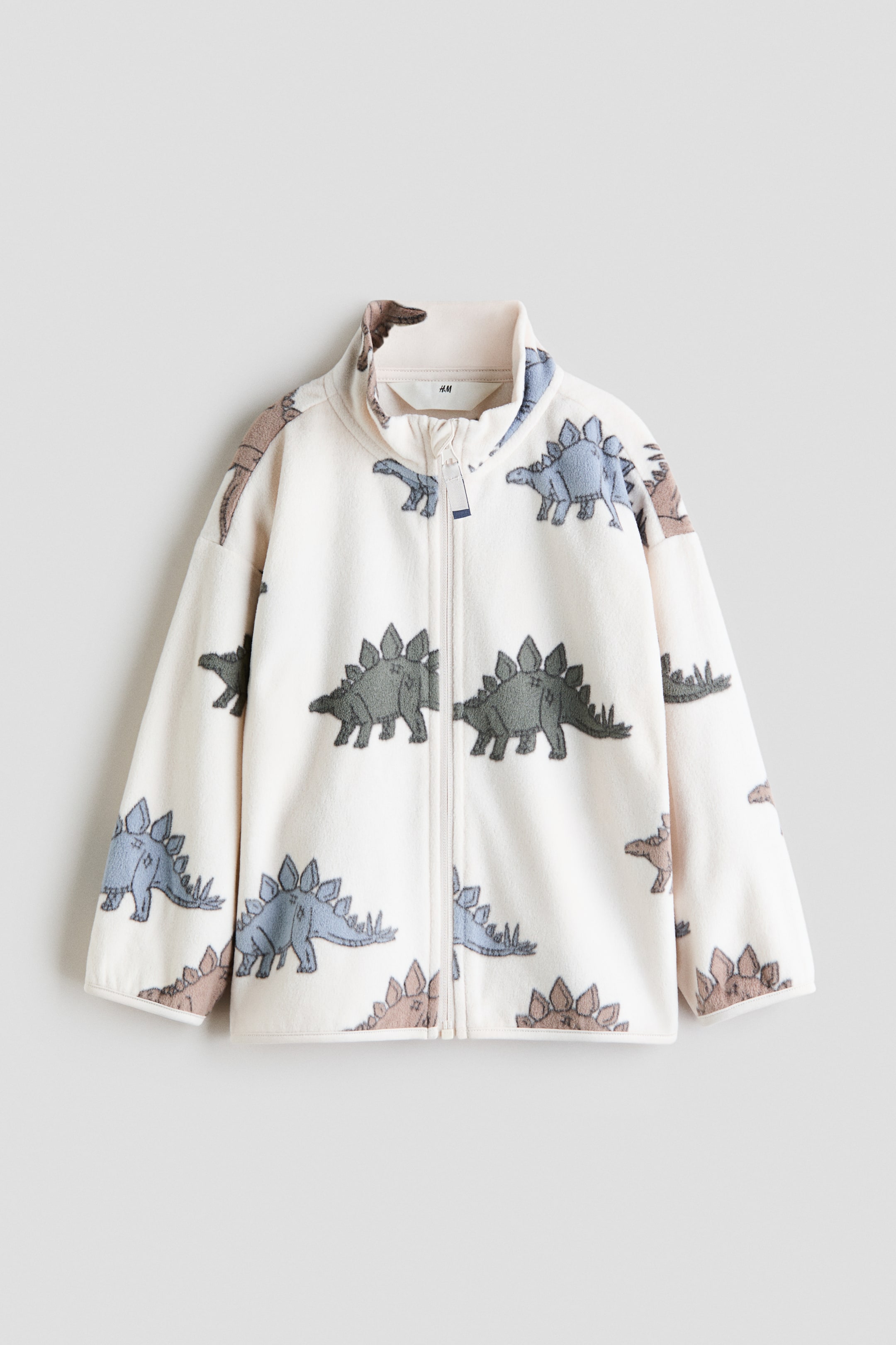 Ampliar la imagen: Una chaqueta de forro polar color crema con cierre completo al frente, cuello alto tipo stand-up y un patrón completo de dinosaurios tipo estegosaurio en tonos azul-gris y marrón-gris.