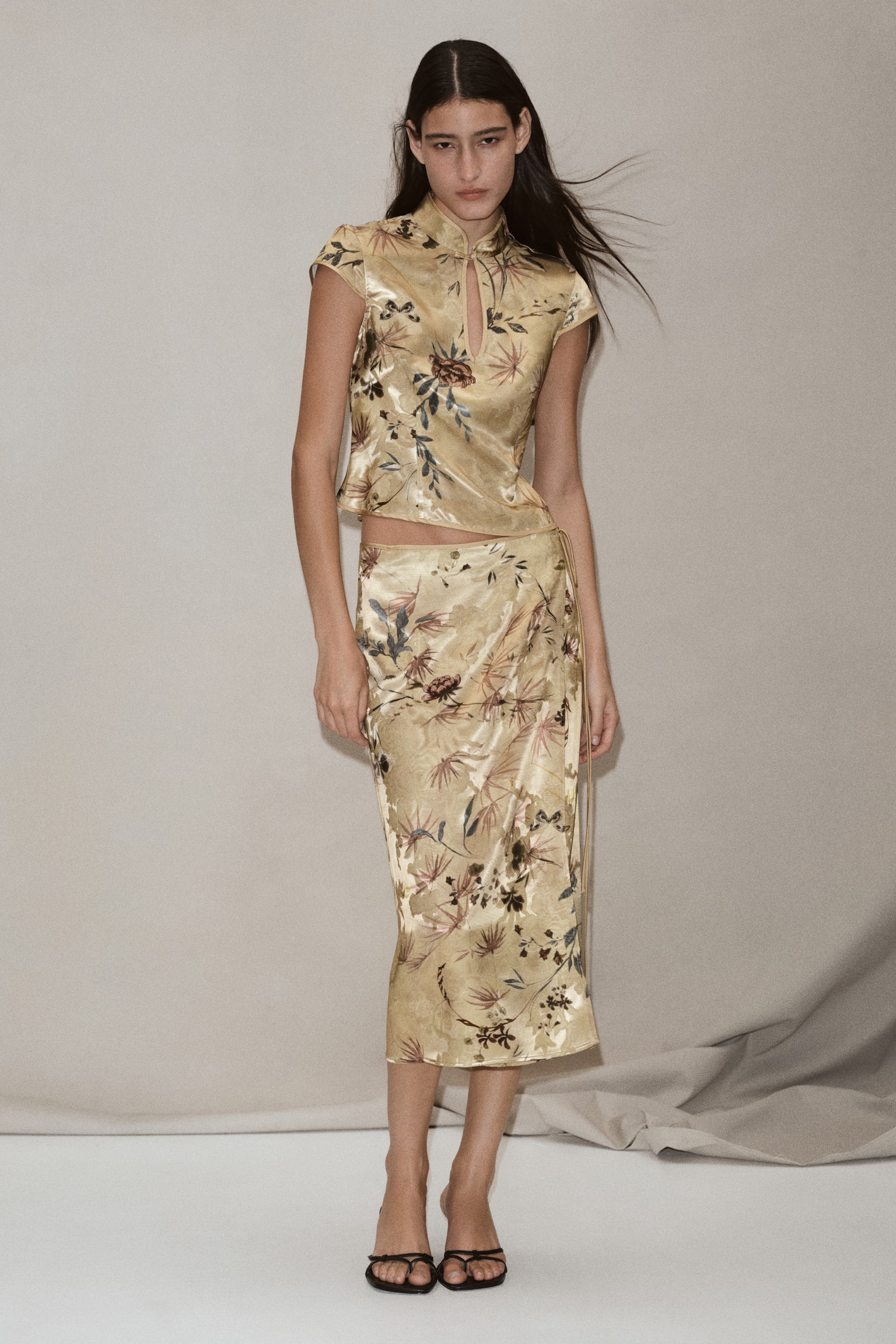 A beige satin wrap skirt is hanging flat, facing front, featuring an all-over jacquard texture beneath a printed botanical motif of pink palm leaves, flowers, and birds. The skirt is shown in a straight silhouette with a delicate vertical tie detail running down the right side. - 1