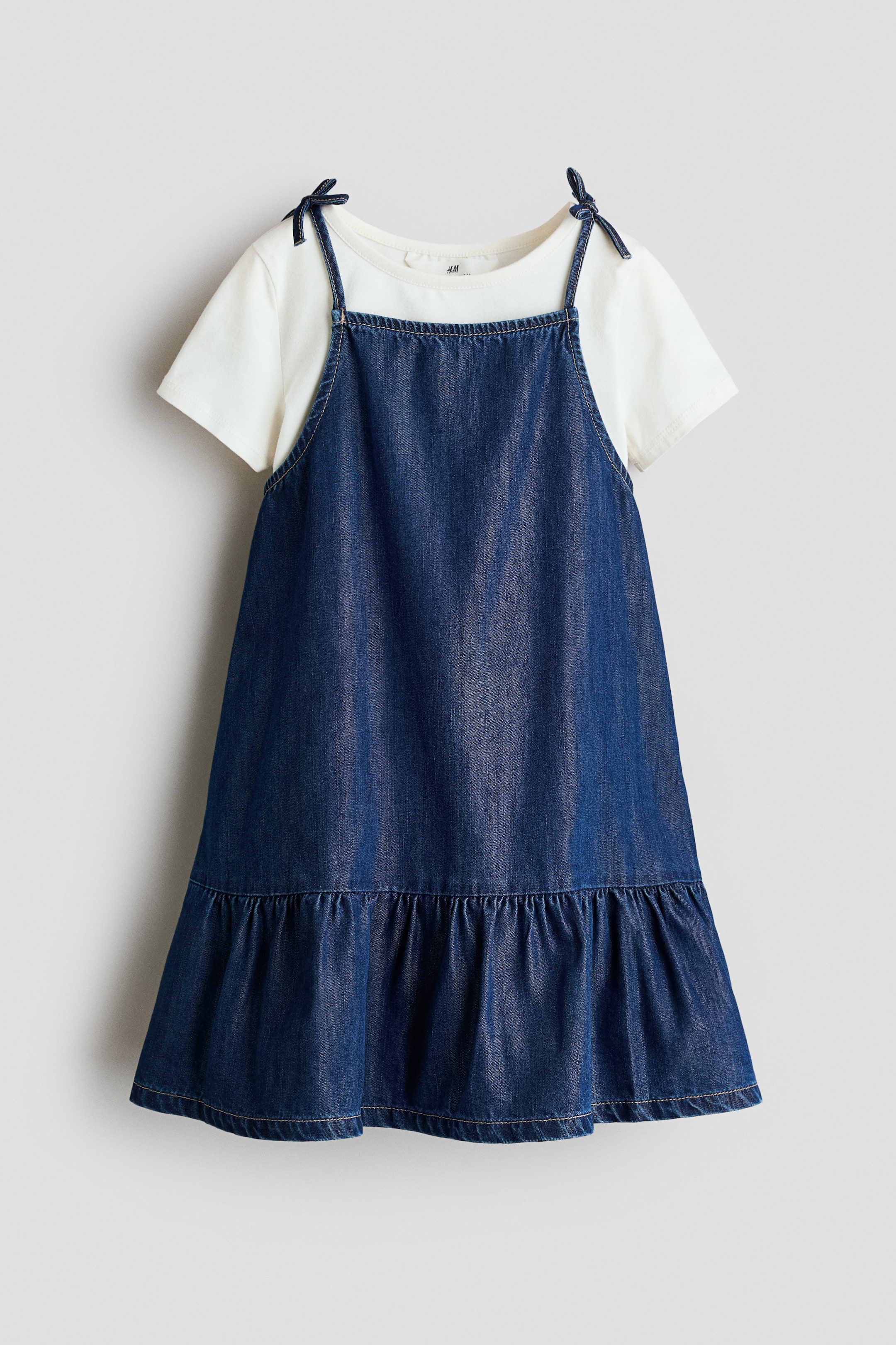 View larger image: A dark blue denim pinafore dress is showcased from the front, featuring tie straps and a ruffled hem. It is styled over a white short-sleeve t-shirt with a round neckline.