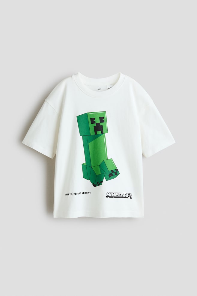 An oversized, off-white t-shirt is presenting its front, made from soft cotton jersey and featuring a large, green, pixelated Minecraft Creeper character printed centrally. The t-shirt is also displaying the text 'CREATE, EXPLORE, SURVIVE' and 'MINECRAFT' below the character, with ribbed trim around its crew neckline and dropped shoulders. - 1
