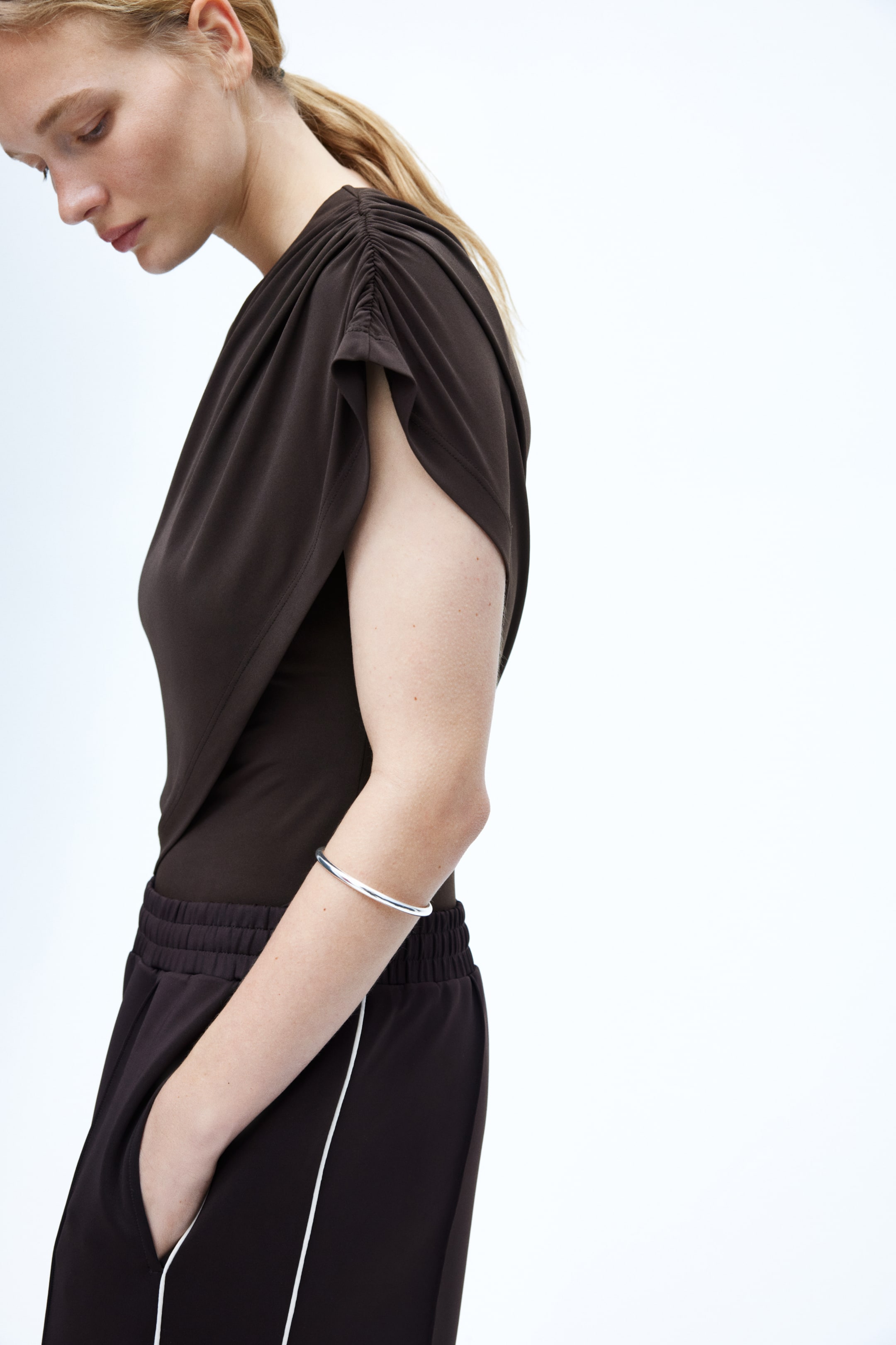 View larger image: A dark brown draped top featuring ruched detailing on the shoulder and flowing cap sleeves. It is paired with matching dark brown track pants, displaying an elasticated waistband, side pockets, and white vertical stripes along the outer seams.
