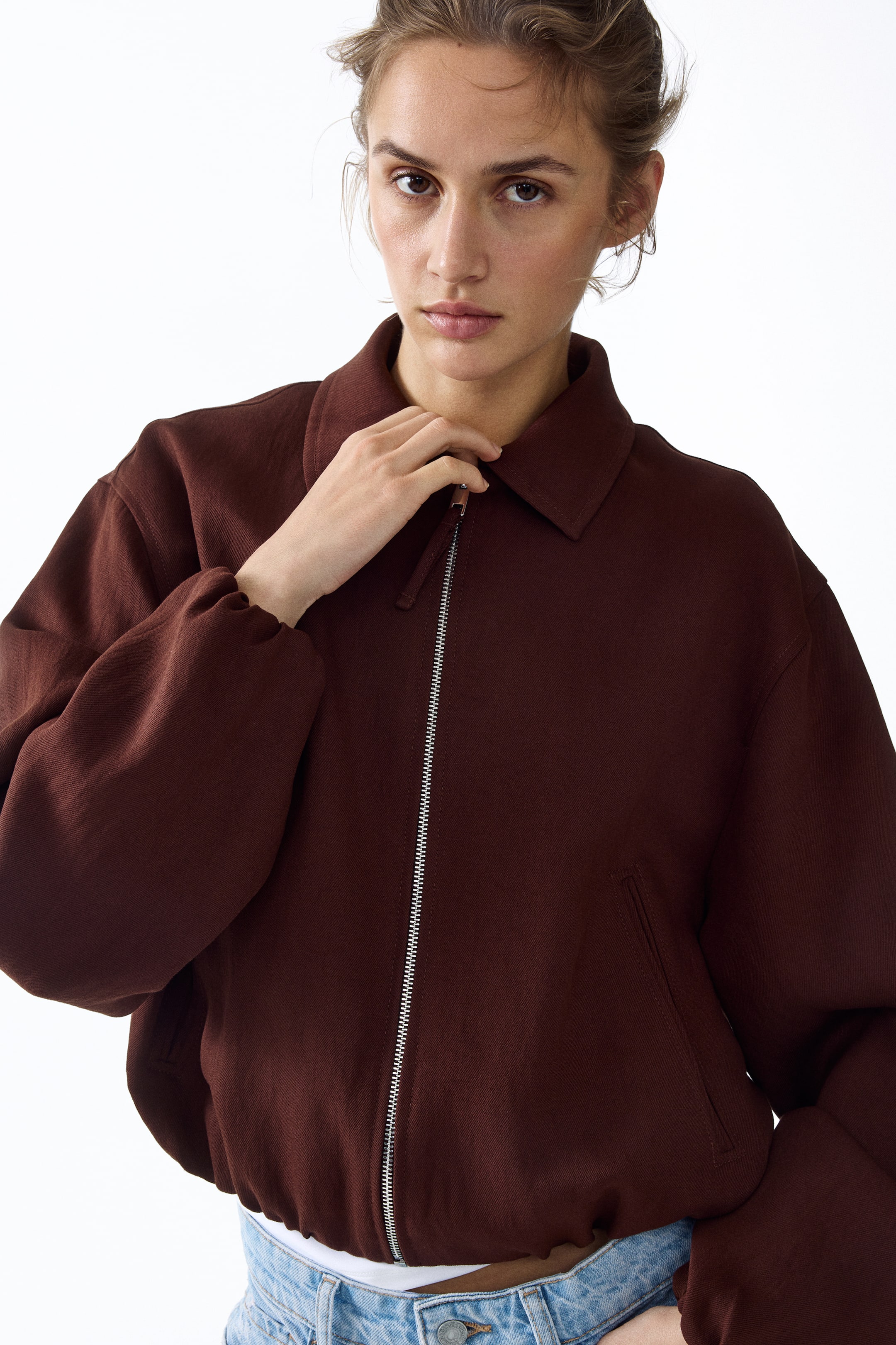 Se større billede: En dyb maroon bomberjakke med en klassisk spids krave og en sølvfarvet frontlynlås, som en hånd holder. Den er designet med voluminøse ærmer, der afsluttes i elastiske manchetter og en elastisk kant, hvilket giver en kort pasform over synlige jeans.