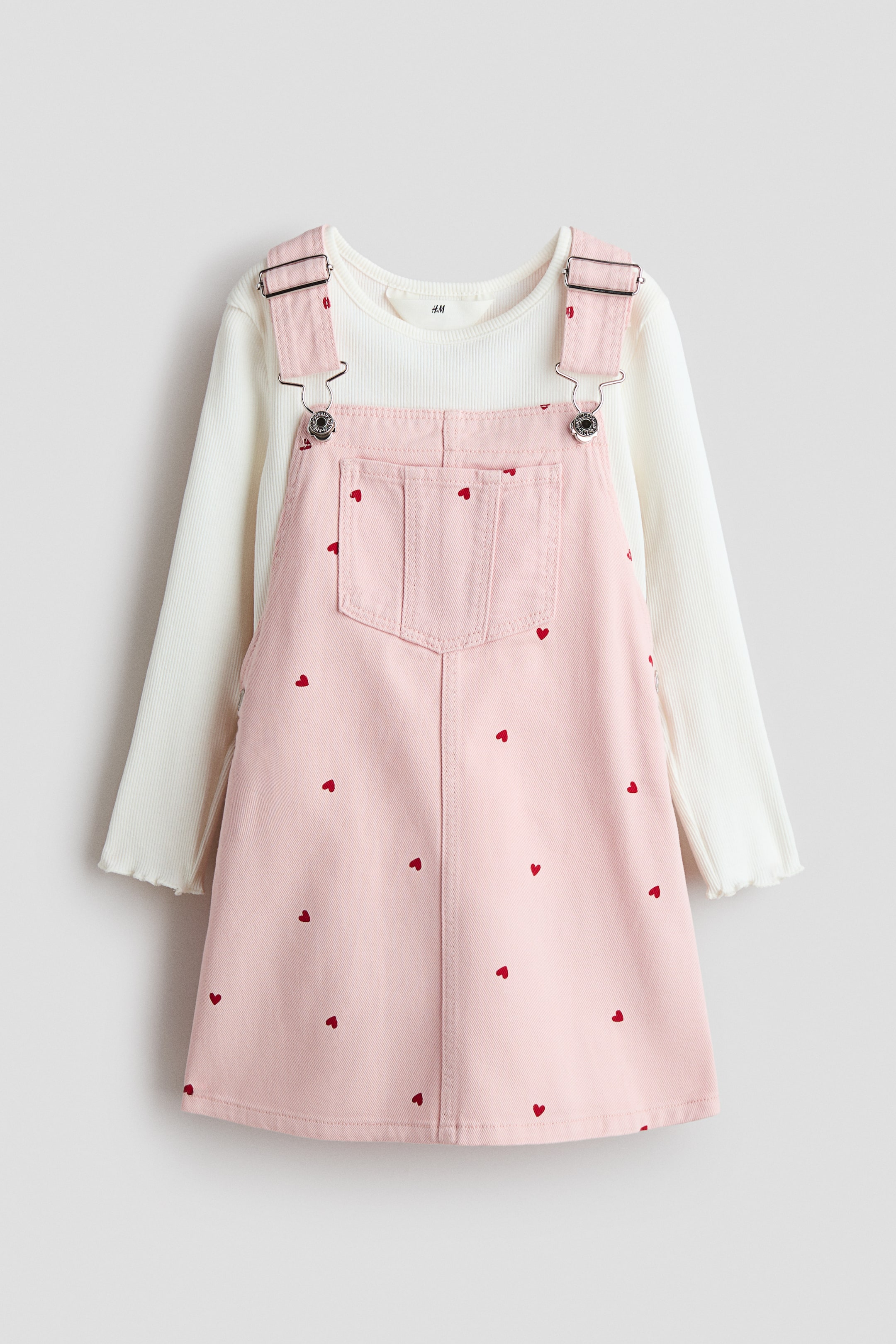 Agrandir l'image: Un ensemble en coton présenté de face met en avant un haut en jersey côtelé doux couleur crème et une robe-salopette en denim rose ornée de motifs de cœurs rouges. Le haut est doté d'un col rond, de manches longues et de bords surjetés, tandis que la robe-salopette présente des bretelles réglables avec attaches métalliques, des pressions sur les côtés et une poche poitrine.