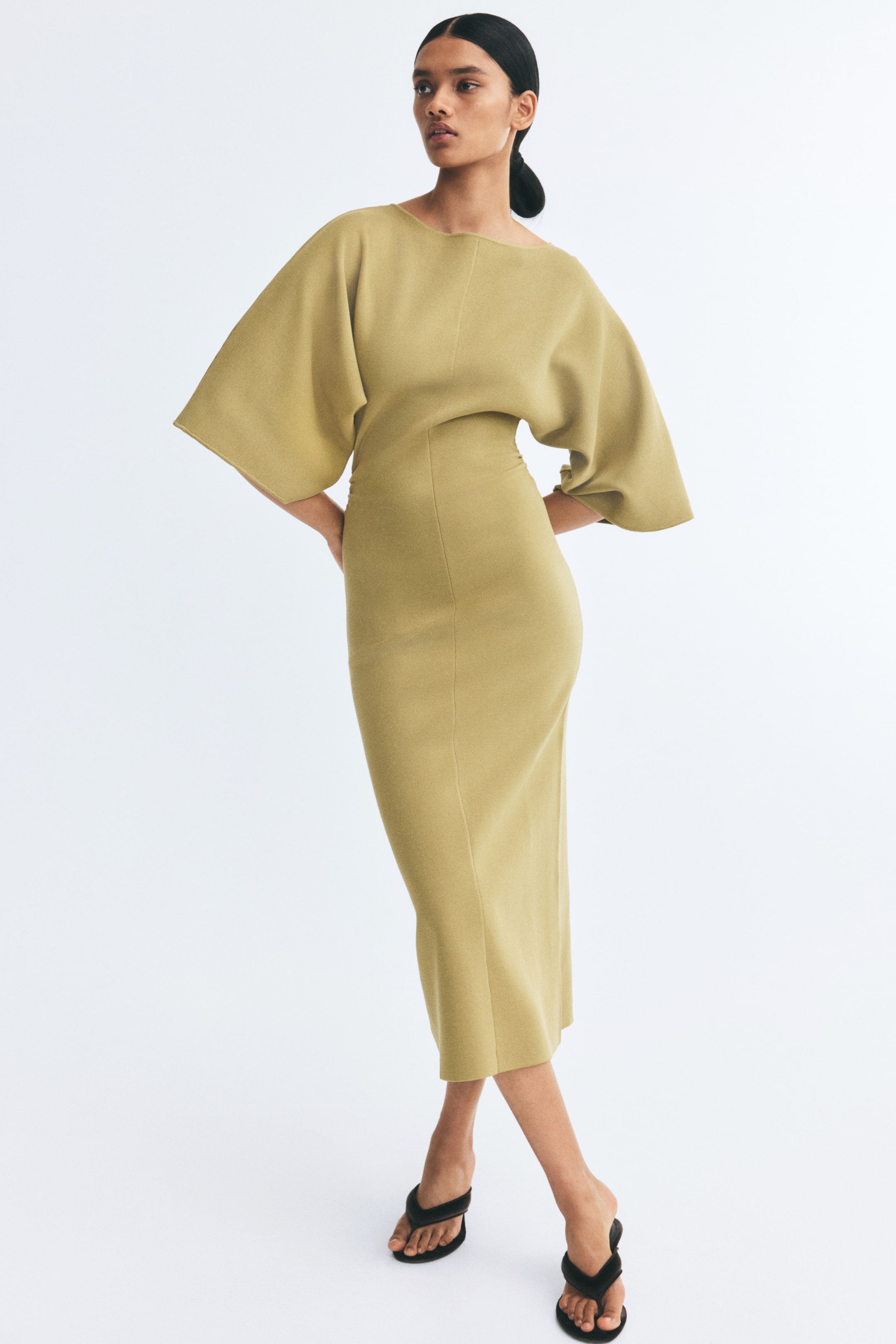 View larger image: A calf-length fitted knit dress in a light olive-yellow colour is being worn by the model, featuring a delicate boat neckline and wide batwing sleeves. The garment is styled with a visible centre seam down the front and gathered side seams that create a draped effect at the waist.