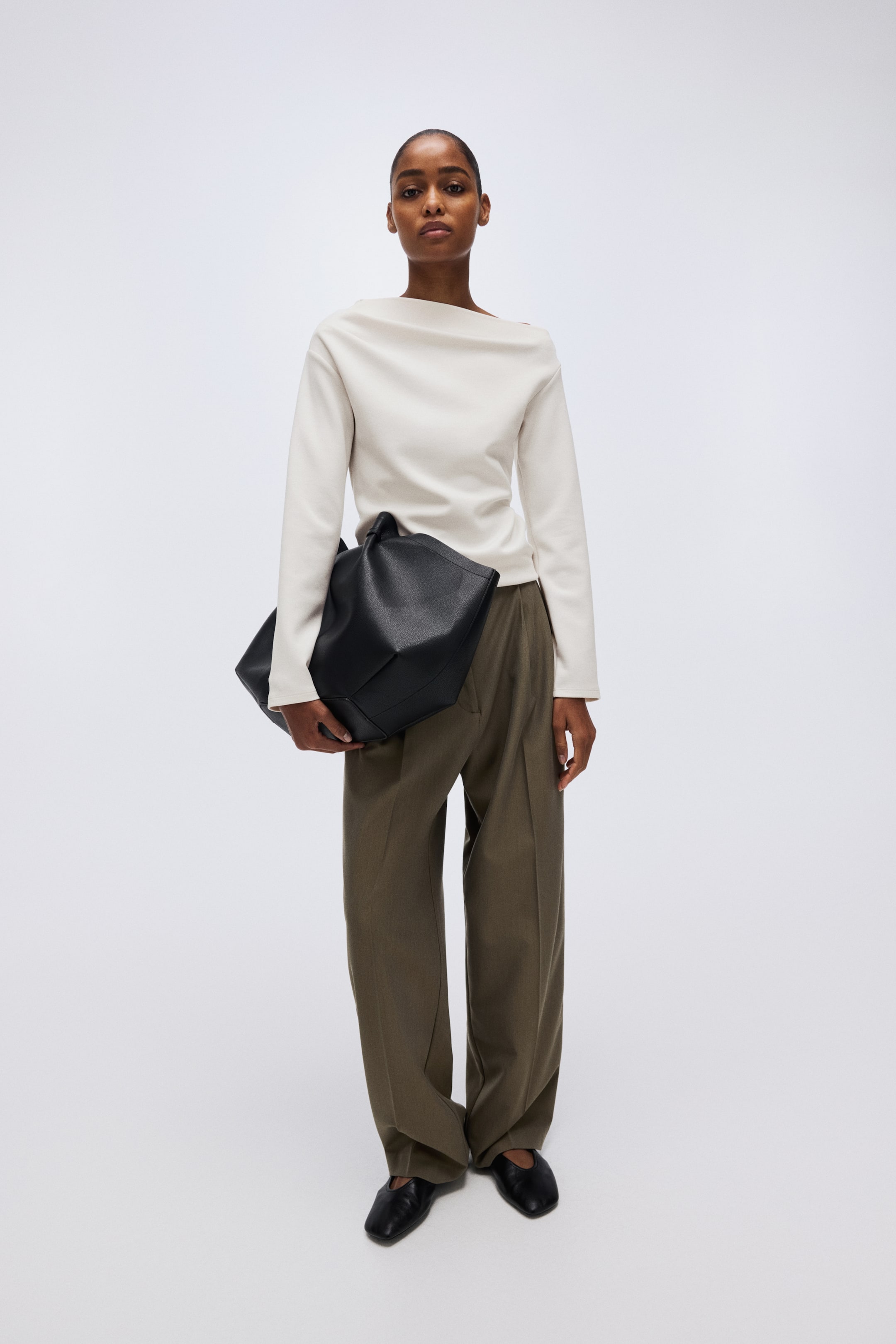 拡大画像を表示: クリーム色の長袖トップスはドレープの効いたワイドネックラインを見せ、オリーブグリーンのワイドレッグプリーツパンツと黒いフラットシューズとコーディネートされ、結び目の付いたハンドルを備えた大きな黒のソフトストラクチャードハンドバッグも合わせて提案されています。