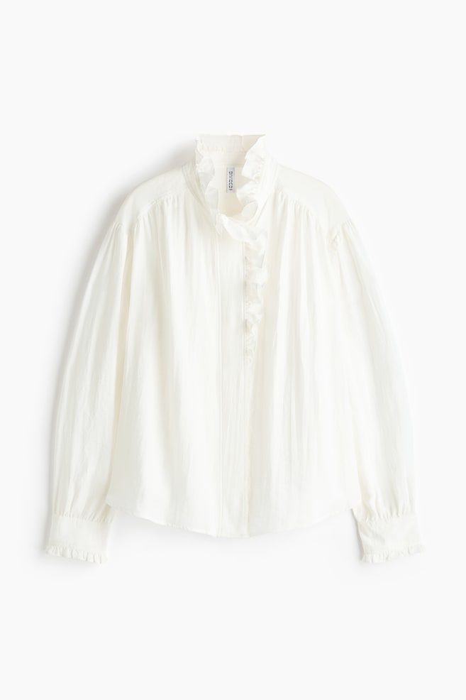Een witte geweven blouse is van voren afgebeeld en laat een hoge ruches kraag zien die overgaat in een verfijnde ruchebies langs één zijde van de blinde knoopsluiting aan de voorkant. De blouse toont lange, volumineuze mouwen met verlaagde schoudernaden en met ruches afgewerkte knoopmanchetten, en eindigt met een zacht afgeronde zoom. - 1