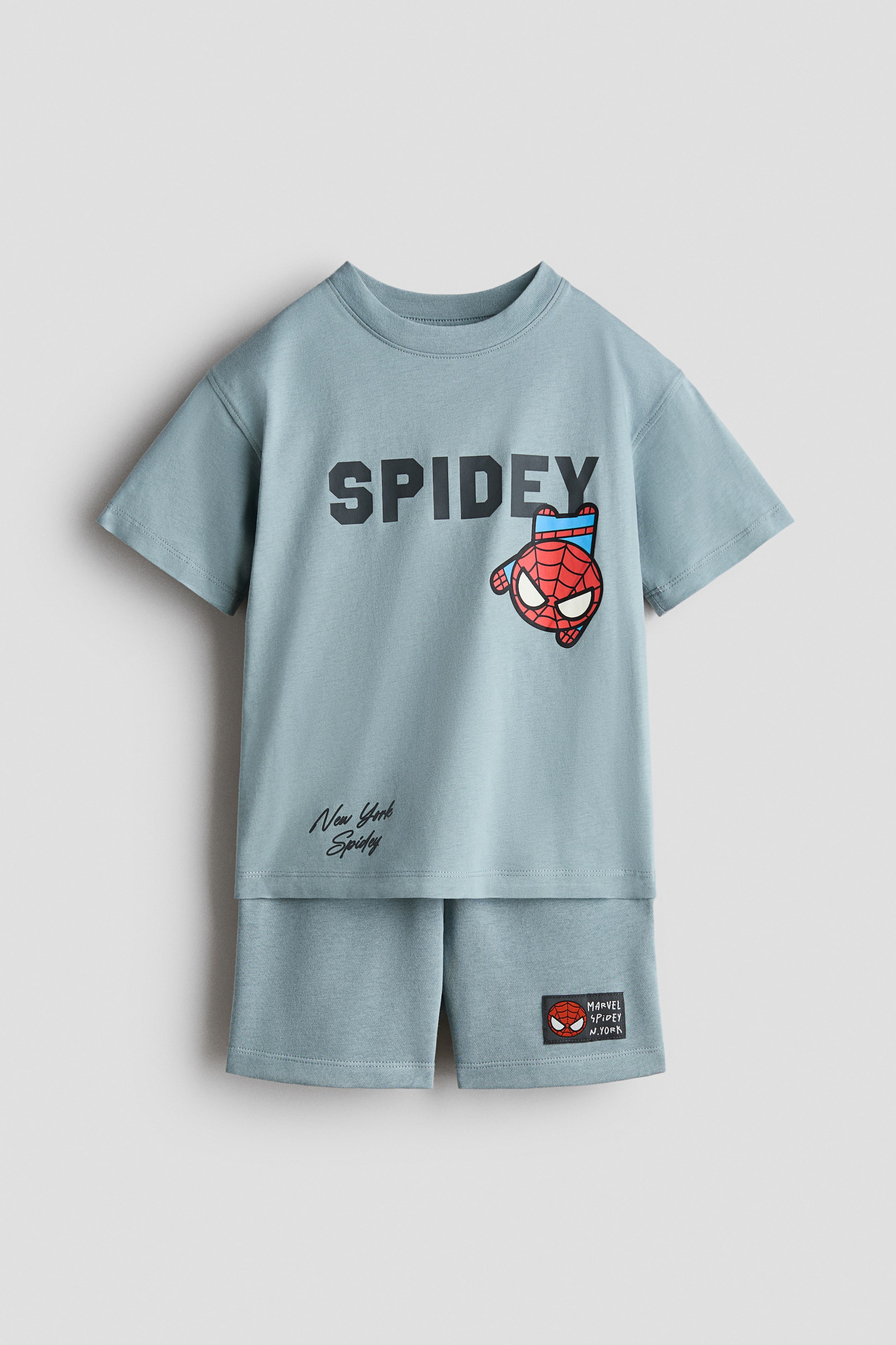 Visa större bild: Ett matchande ljusblått T-shirt och shorts set, tillverkat av mjukt tyg, visas framifrån. T-shirten i bomullsjersey har "SPIDEY" i svarta bokstäver och ett Spider-Man-huvudgrafik på bröstet, med "New York Spidey" skrivet nedanför, och är designad med nedhasade axlar, vida ärmar och en ribbad halsringning; samtidigt har de lätta shorts i sweatshirt-tyg en liten patch med ett Spider-Man-huvud och "MARVEL SPIDEY N.YORK" på vänster ben, och är designade med en täckt elastisk midja.