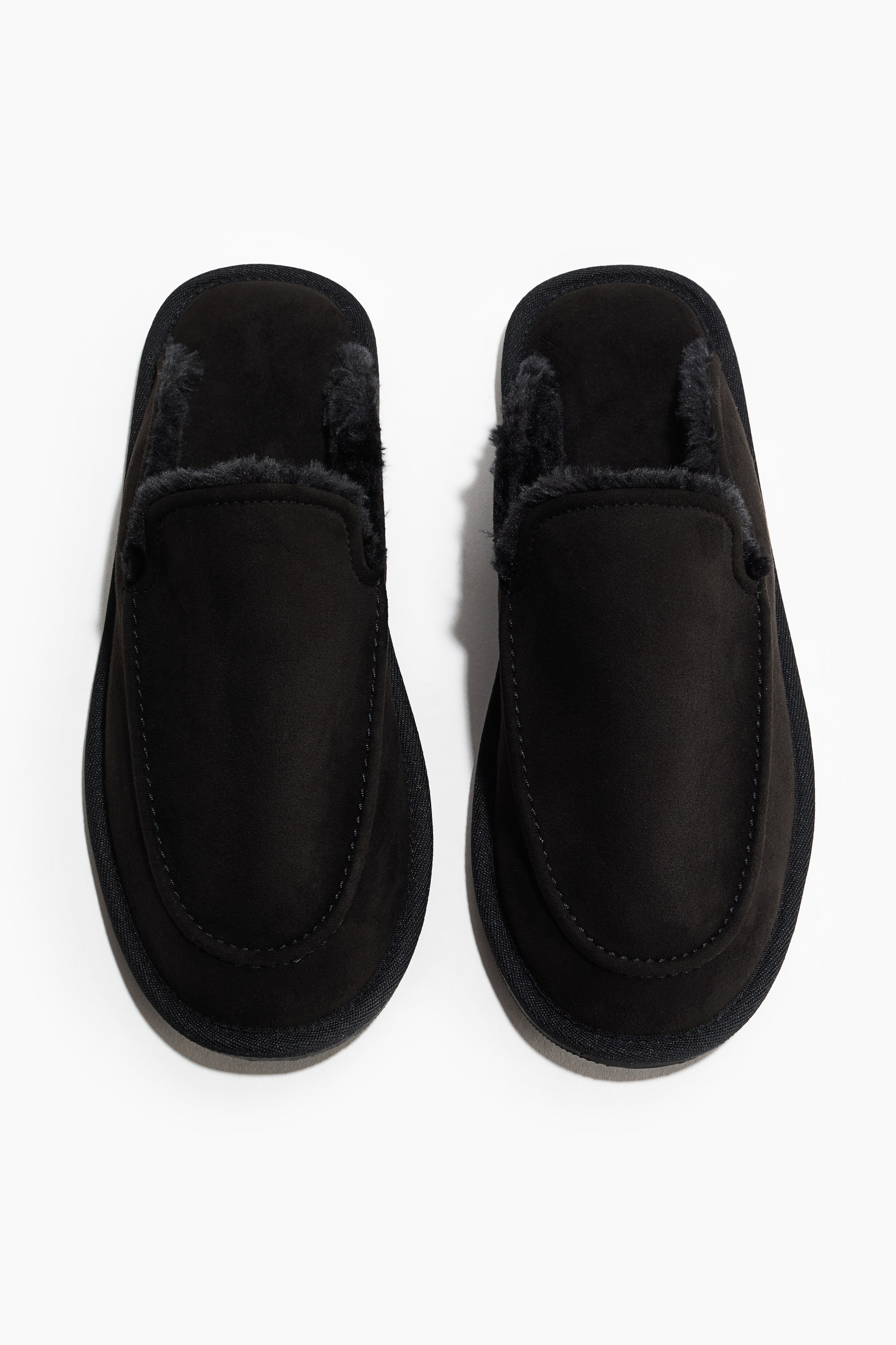 Agrandir l'image: Une paire de pantoufles noires présente une tige effet suédé avec une doublure en fausse fourrure foncée visible autour de l’ouverture et un bout cousu de style mocassin. Les pantoufles sont orientées vers l’avant, vues directement du dessus.