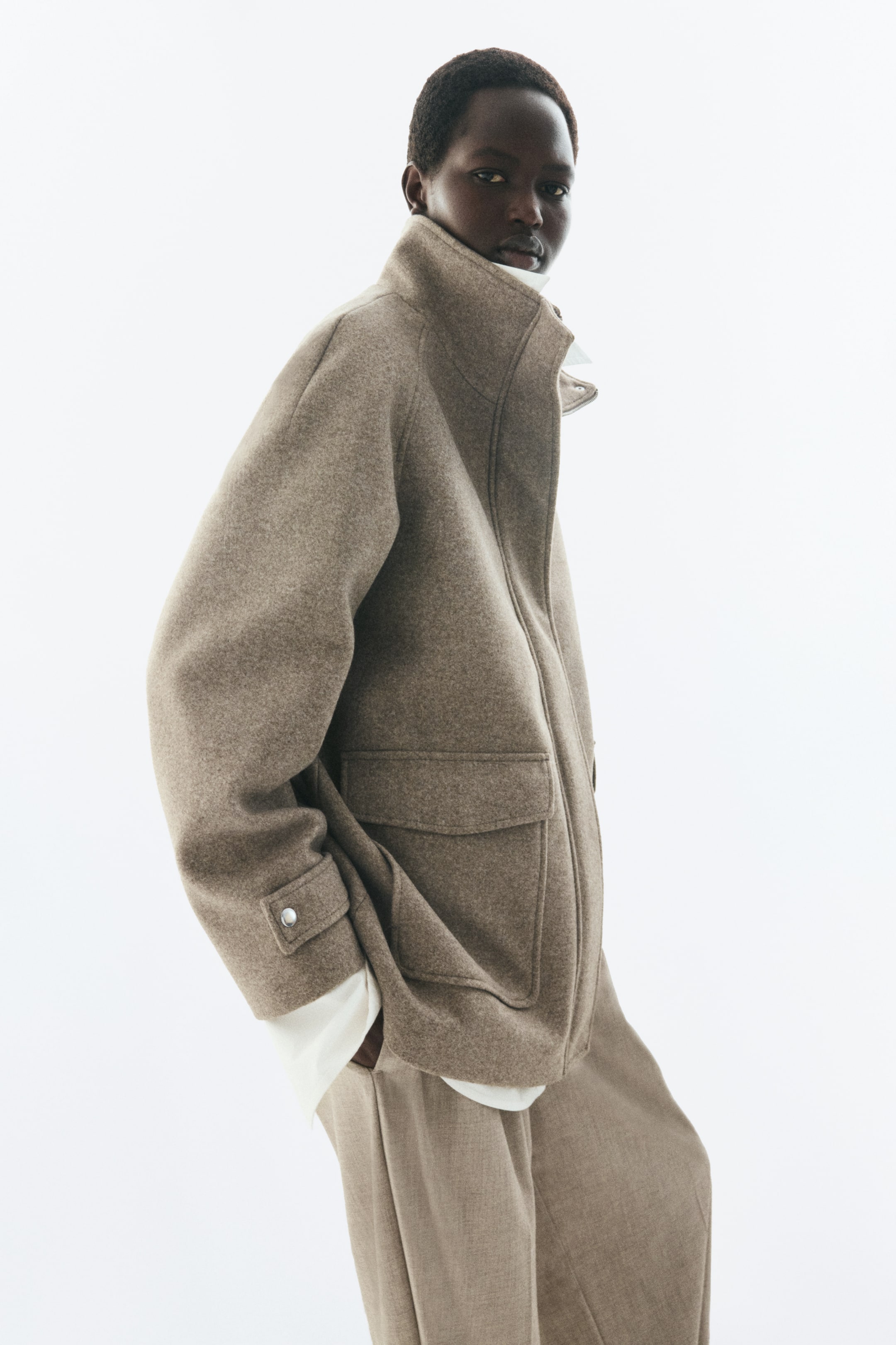Voir une image plus grande: Un manteau beige en tissu feutré est porté par le mannequin, doté d’un col cheminée montant, de manches raglan avec pattes de poignet à boutons-pression, et d’une poche plaquée à rabat sur le devant. Le manteau est superposé sur une chemise blanche et agencé avec un pantalon beige assorti.