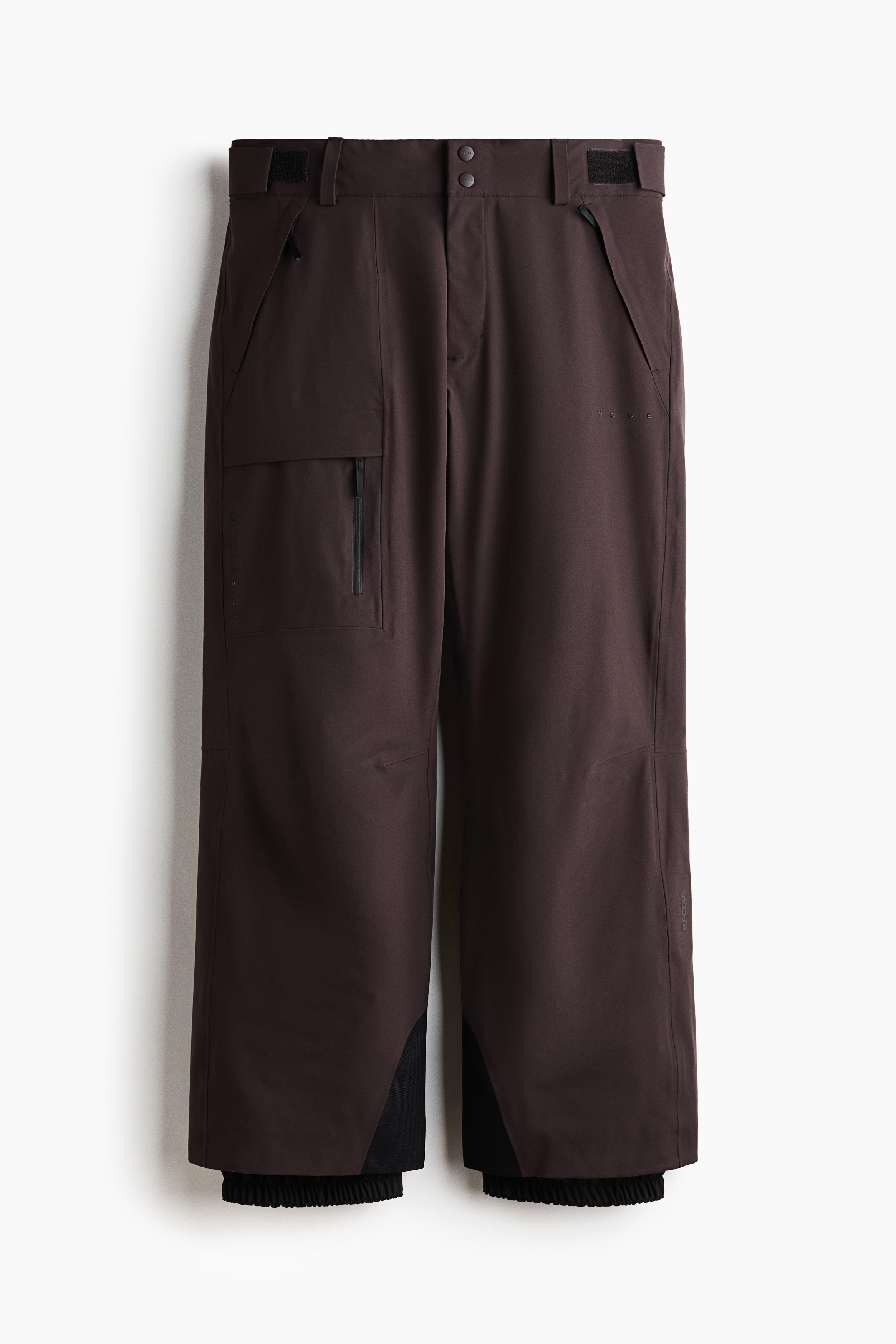 Ein dunkelbrauner technischer Outdoor-Anzug, bestehend aus einer Kapuzen- ReiÃverschlussjacke und einer passenden locker geschnittenen Hose, wird vom Model getragen. Das Model steht mit den HÃ¤nden in den Hosentaschen, wobei die Hose Ã¼ber schwarzen Skistiefeln getragen wird.