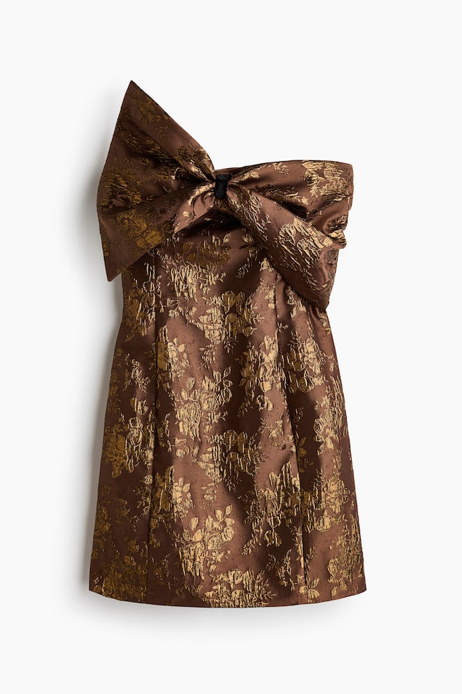 Olkaimeton minimekko on kuvattu edestÃ¤, ja siinÃ¤ on ruskea ja metallinhohtoinen kullanvÃ¤rinen kukkakuvioinen jacquard-pinta. Mekossa on suuri, rakenteellinen ja epÃ¤symmetrinen rusetti, joka laskeutuu ylÃ¤osan yli, sekÃ¤ hieman levenevÃ¤ hame, jossa on pystysuuntaiset muotosaumat. - 1