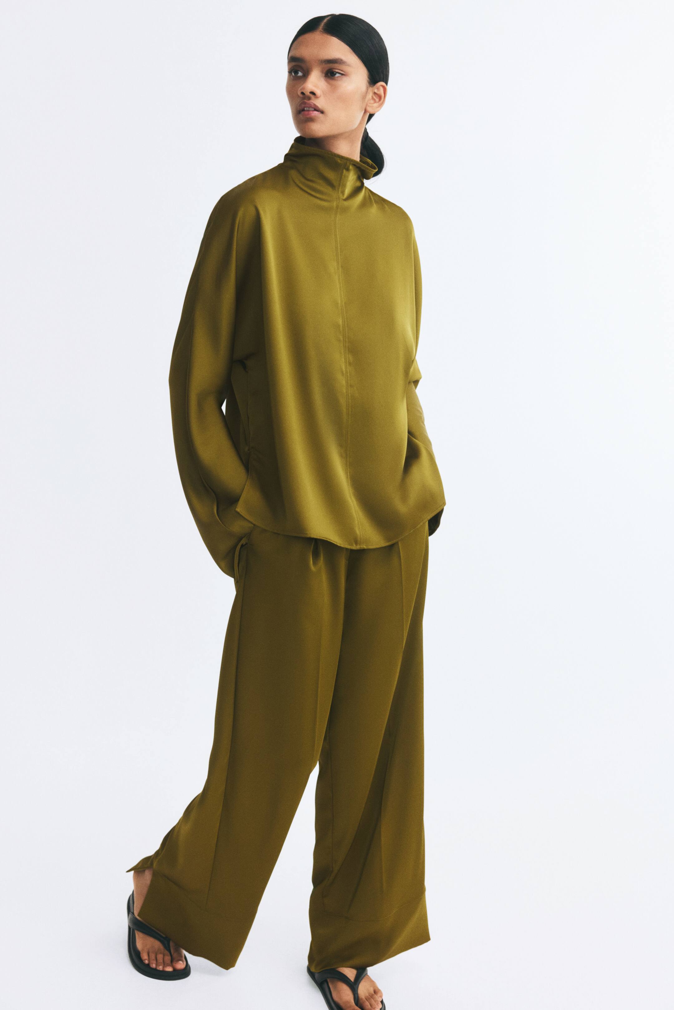 Agrandir l'image: Le mannequin porte un pantalon en satin vert olive à la coupe ample et à jambes droites, avec des plis sur le devant. Le pantalon est associé à un haut en satin assorti à col montant et à des sandales plates noires.