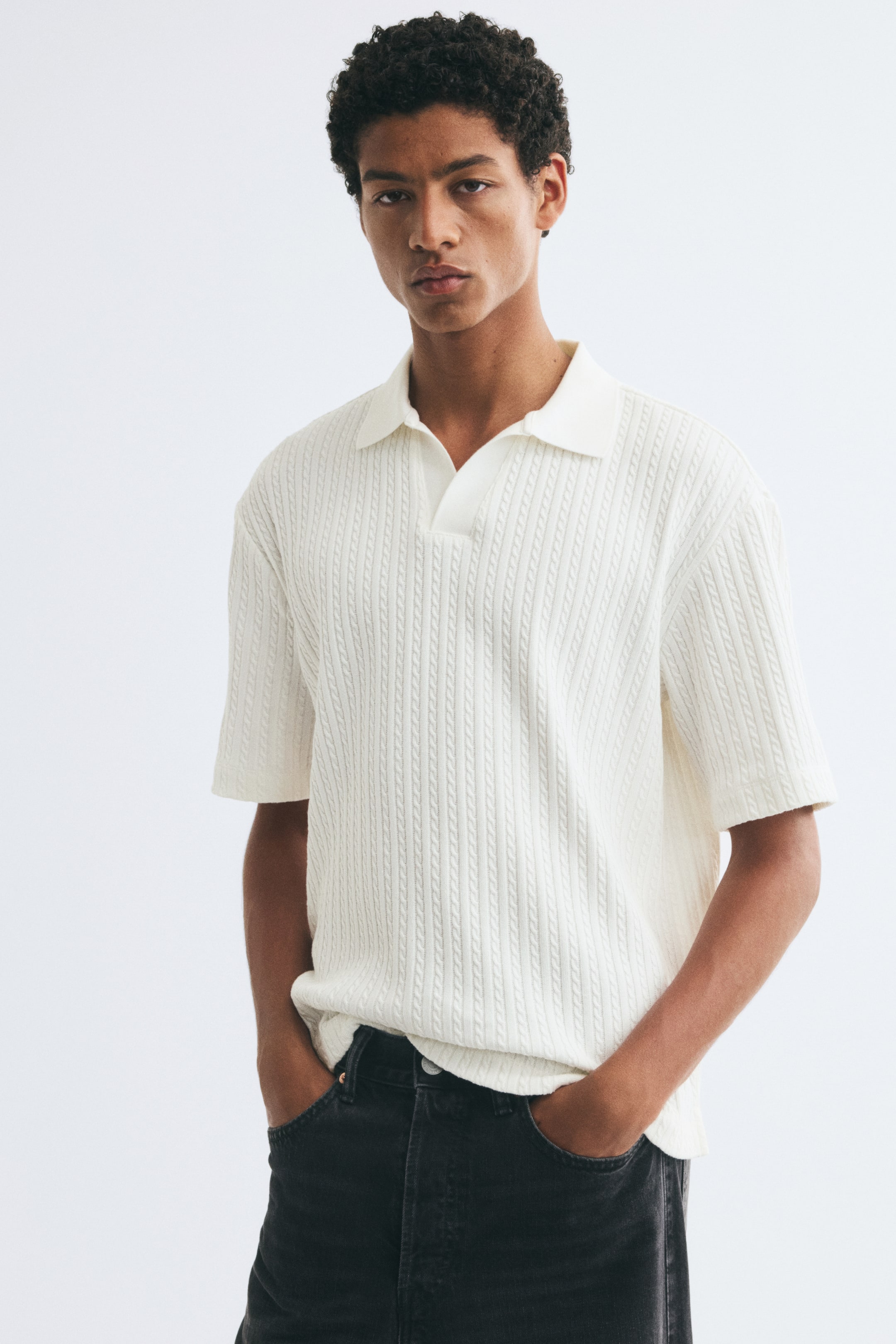 View larger image: The model is wearing a cream-coloured, short-sleeved polo shirt in a vertical cable-knit pattern. The shirt features an open collar and a relaxed fit, and is styled with dark grey jeans as the model stands with hands in the pockets.