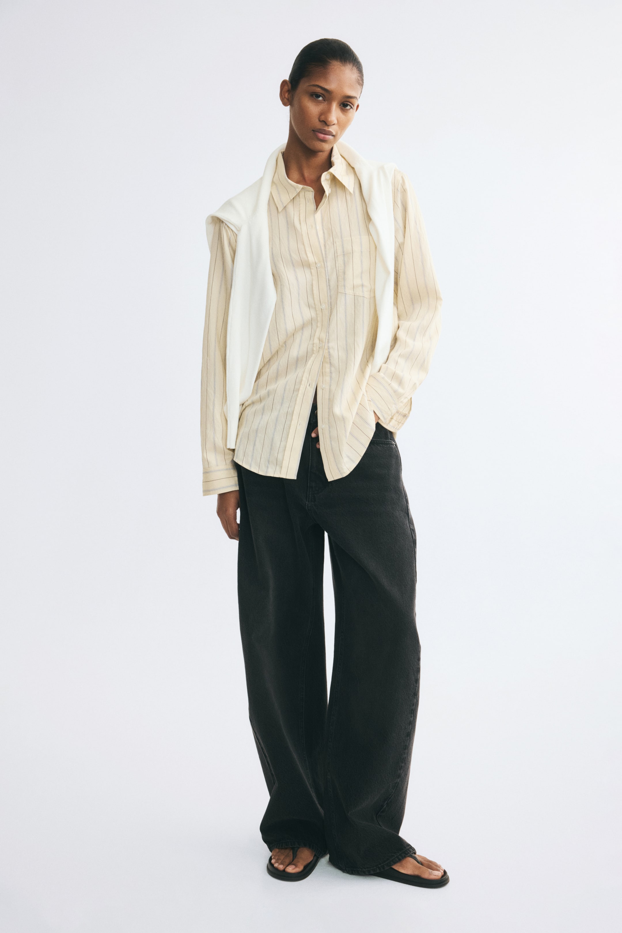 View larger image: A light yellow, vertically striped button-down shirt is worn by the model, paired with wide-leg dark jeans and a white sweater draped over the shoulders. The long-sleeved shirt features a collar, a front button closure, and a rounded hem, with the model standing with one hand tucked into a pocket.