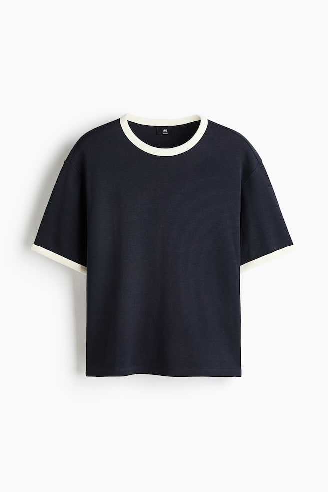 Una camiseta de jersey azul oscuro muestra su parte delantera, con un cuello redondo ribeteado y dobladillos de manga en un color blanco roto contrastante. Está diseñada con hombros caídos y un dobladillo de corte recto, mostrando un ajuste relajado. - 1