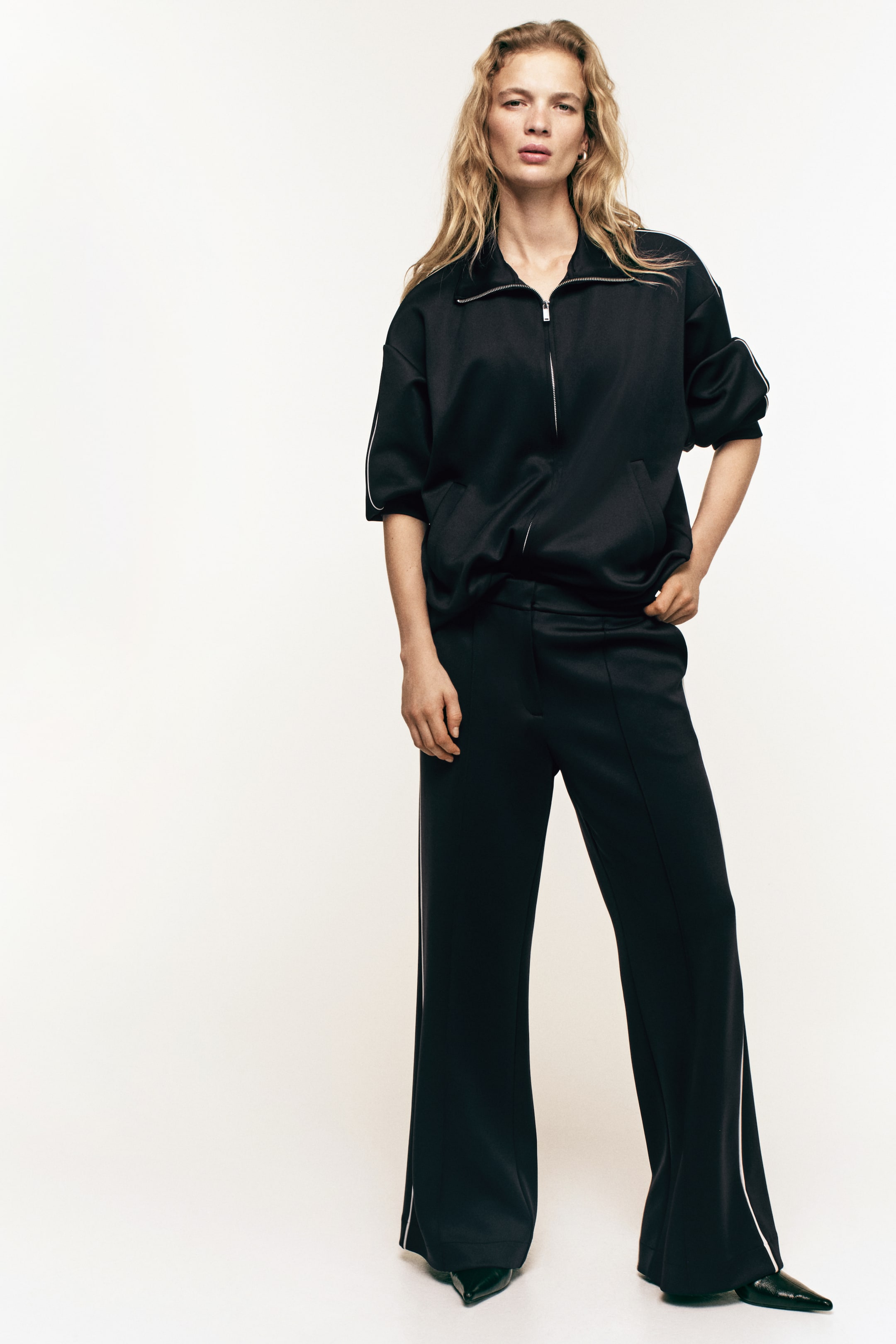 View larger image: Black scuba fabric trousers feature diagonal side pockets, straight legs with sewn-in creases down the front, and accent-colour piping along the side. The model is wearing the trousers, with one hand resting in a side pocket.