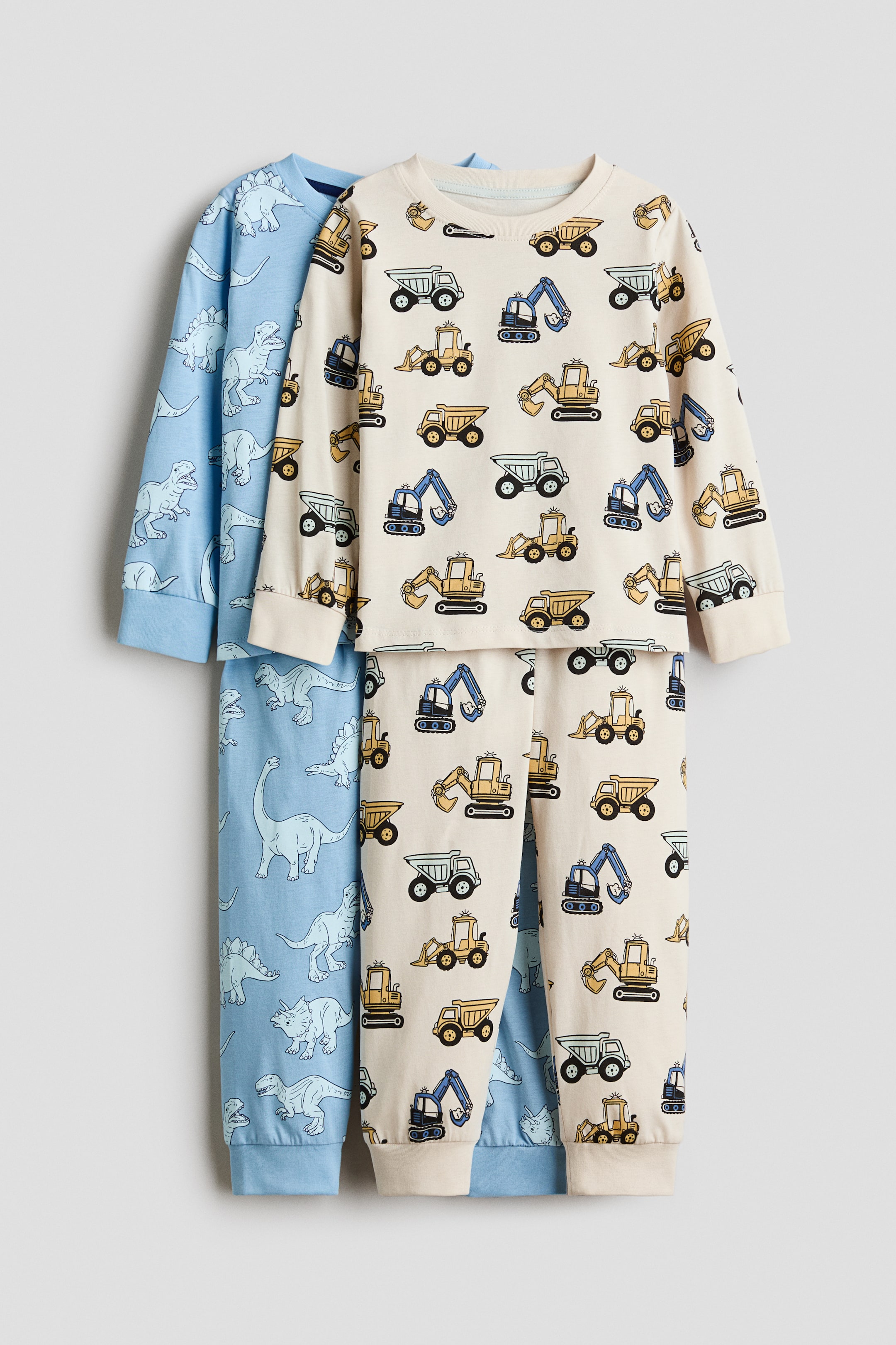 Visualizza l'immagine ingrandita: Due set di pigiami per bambini sono appesi frontalmente, con un set color crema con motivo di veicoli da costruzione sovrapposto a un set azzurro chiaro con motivo di dinosauri. Entrambi i set mostrano maglie a maniche lunghe con scollo rotondo a coste e polsini, abbinate a pantaloni coordinati con fondo gamba a coste.