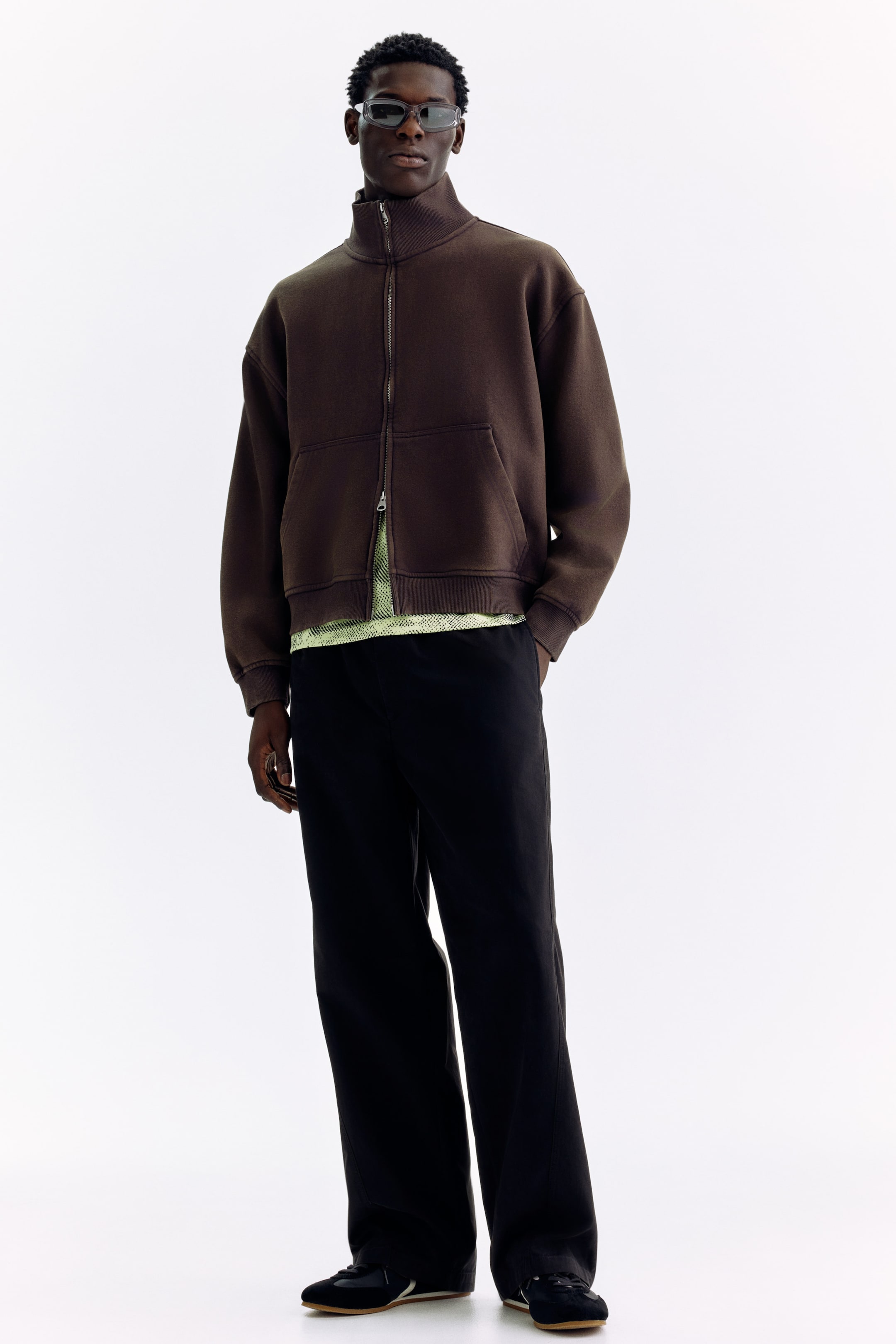 Visa större bild: En mörkbrun jacka med hel dragkedja bärs, med en hög ståkrage, en silverfärgad dragkedja och två stora påsydda fickor vid midjan. En ljusgrön mönstrad skjorta är synlig under dess avslappnade passform och nedhasade axlar.