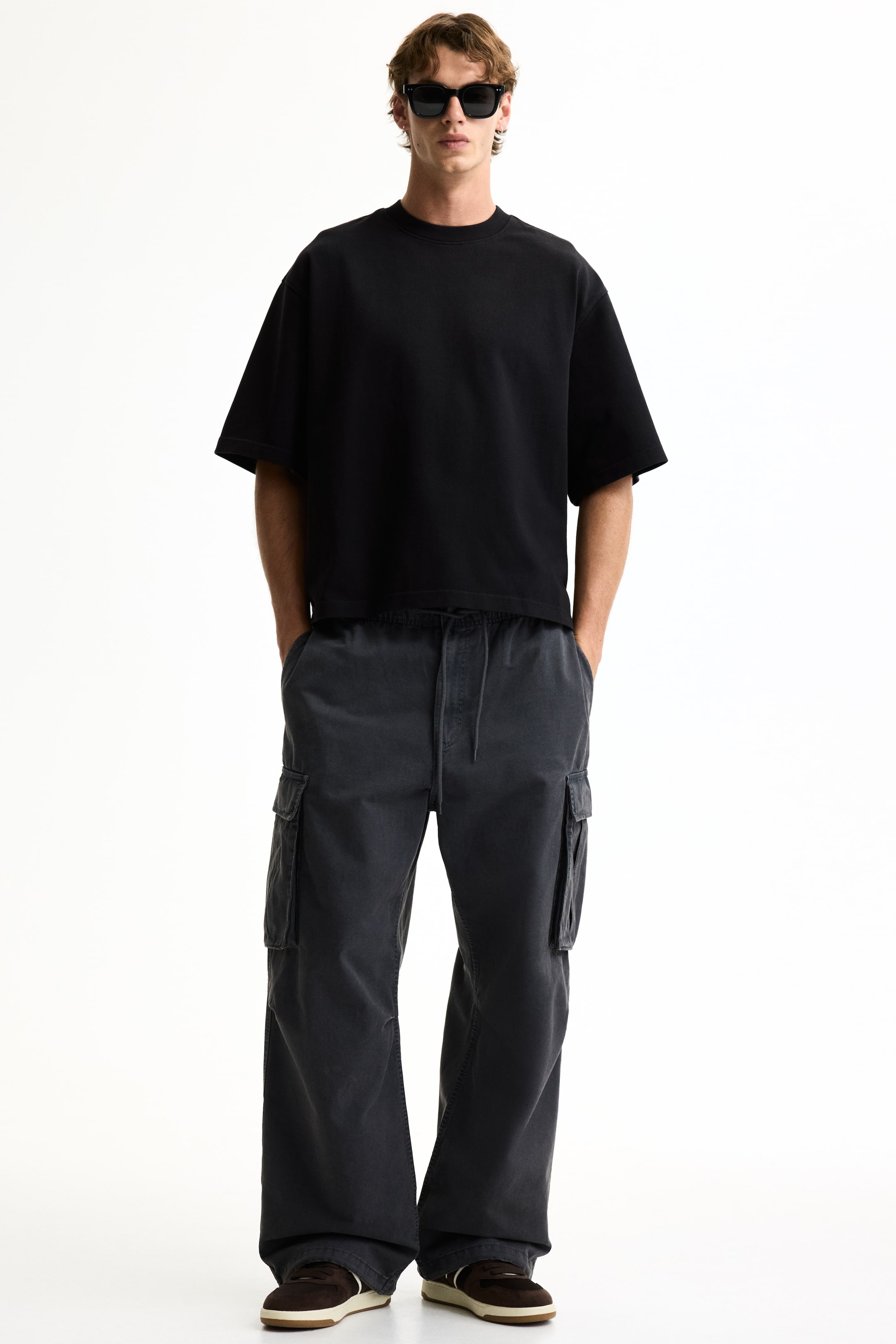Voir une image plus grande: Un t-shirt noir surdimensionné avec un col rond et de larges manches courtes, porté avec un pantalon cargo gris foncé à jambes larges. Le pantalon présente une taille élastique avec cordon de serrage, de grandes poches cargo latérales, et repose sur des baskets marron et blanches.
