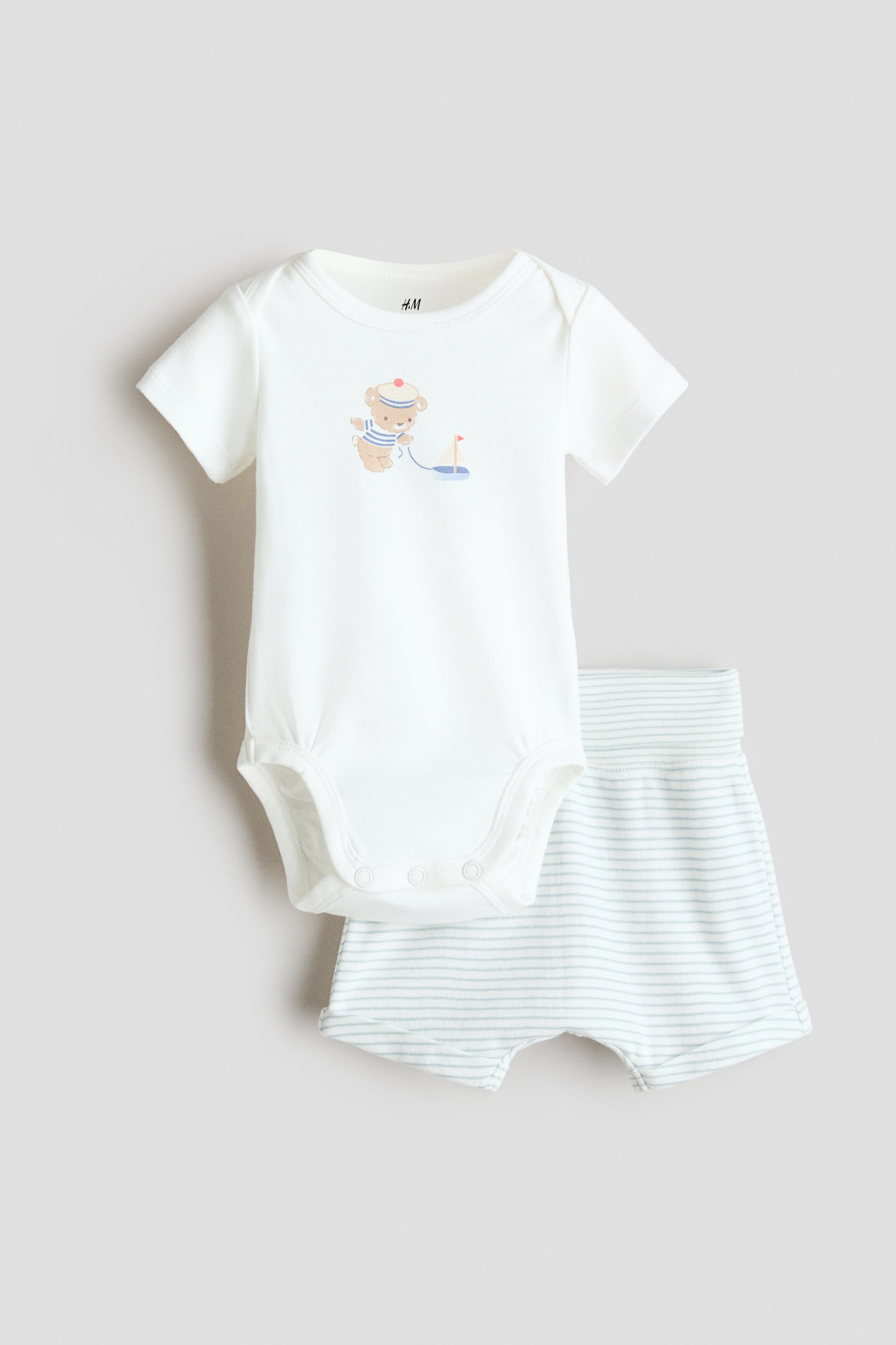 View larger image: A white short-sleeved bodysuit with an envelope neck and a bear graphic pulling a sailboat on the chest is layered over a pair of white and light blue striped shorts, both presented front-facing. The bodysuit features press-studs at the bottom, while the shorts showcase a foldover waistband and sewn-in turn-ups at the hems.