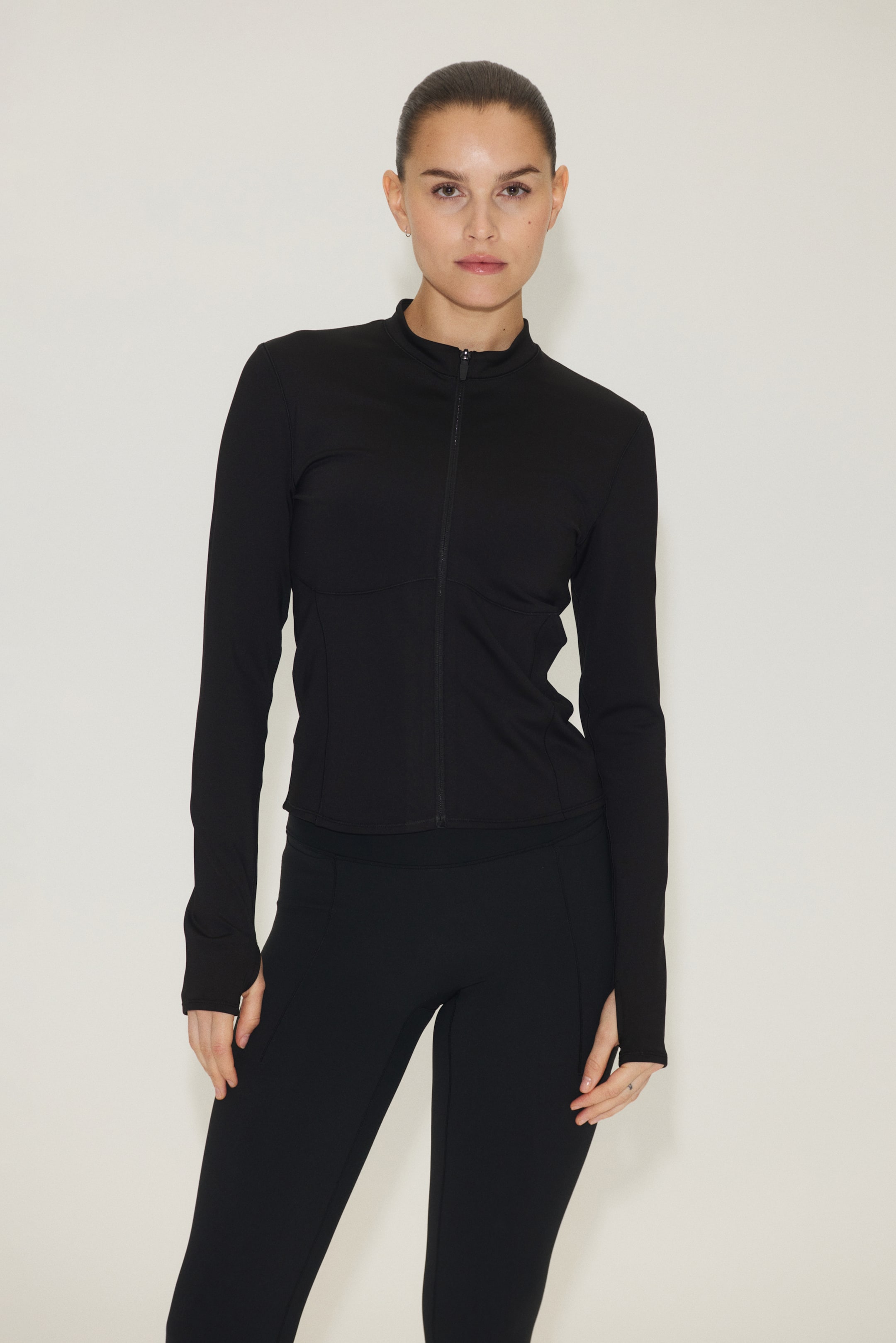 Nagyobb kép megtekintése: Egy fekete sportdzseki látható elölről, amely álló gallérral és teljes hosszában futó elülső cipzárral rendelkezik. Testhez simuló kialakítást mutat be, kontúros varrásvonalakkal és hosszú ujjakkal, amelyek hüvelykujjnyílásokkal vannak ellátva.