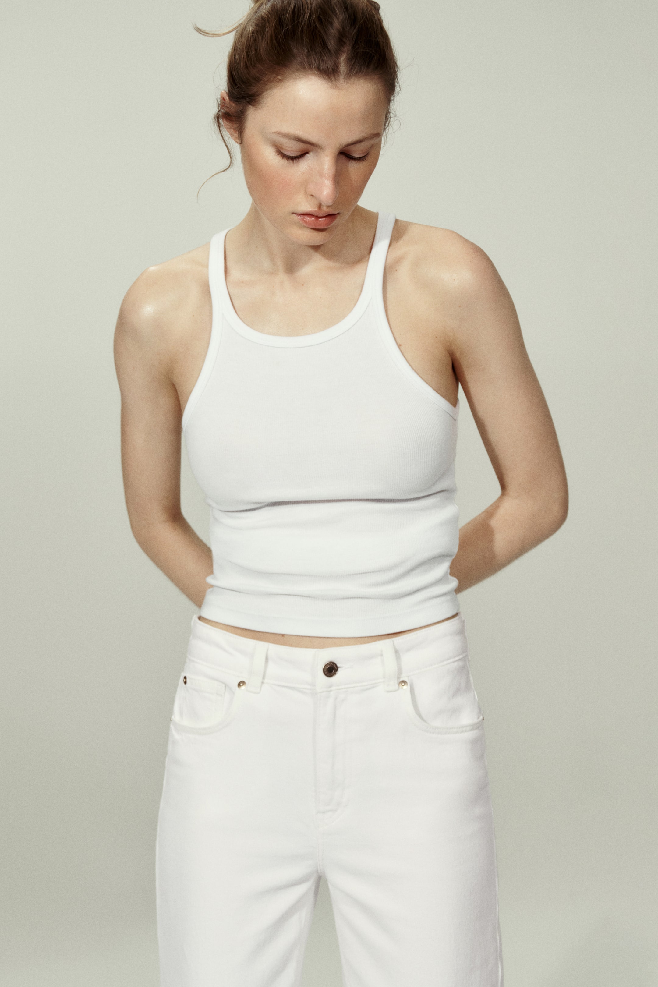 查看大圖: 穿著一件白色羅紋背心，特點是圓領和寬肩帶，搭配一條白色牛仔褲，展示五口袋設計和前扣閉合。