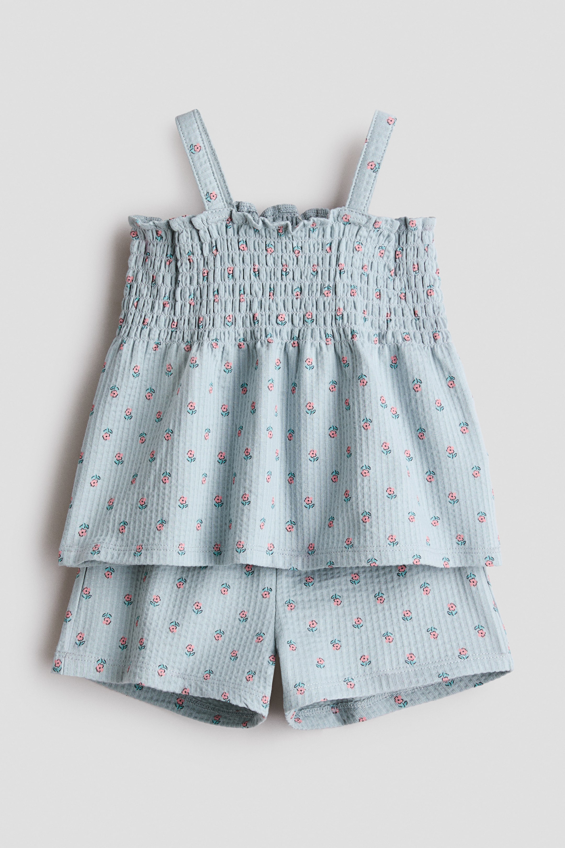 Agrandir l'image: Un ensemble deux pièces bleu clair est présenté de face, composé d'un haut et d'un short. Le haut présente un corsage froncé avec des bretelles et un ourlet évasé, et le short est de longueur courte assortie, tous deux affichant un petit imprimé floral rose et rouge sur un tissu côtelé.