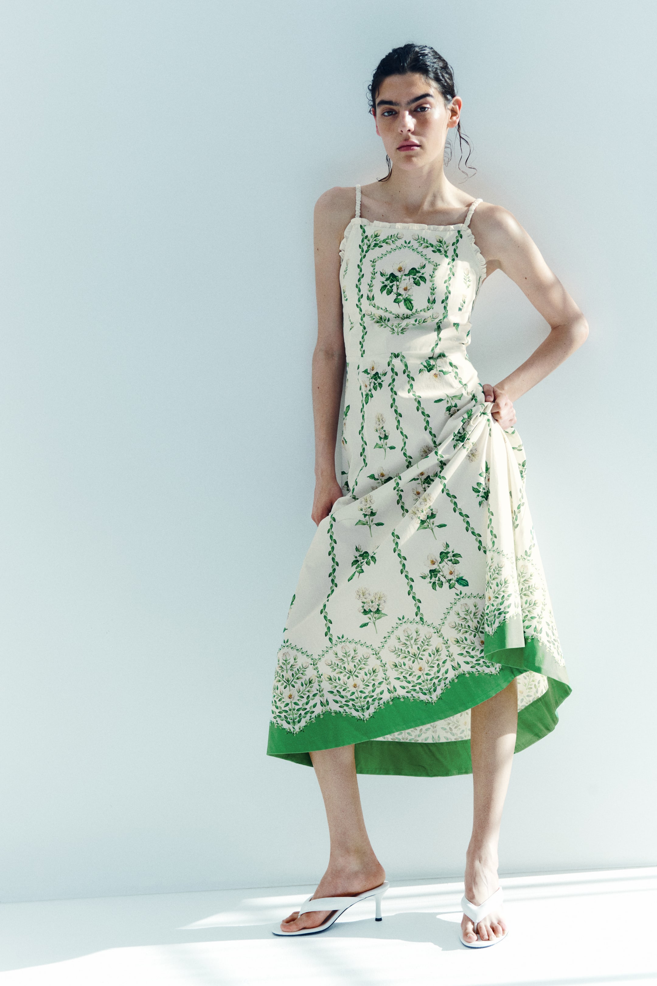 View larger image: An off-white midi dress with spaghetti straps and a ruffled neckline is displayed, showcasing green botanical patterns and a wide green border along its hem. White heeled thong sandals are worn.