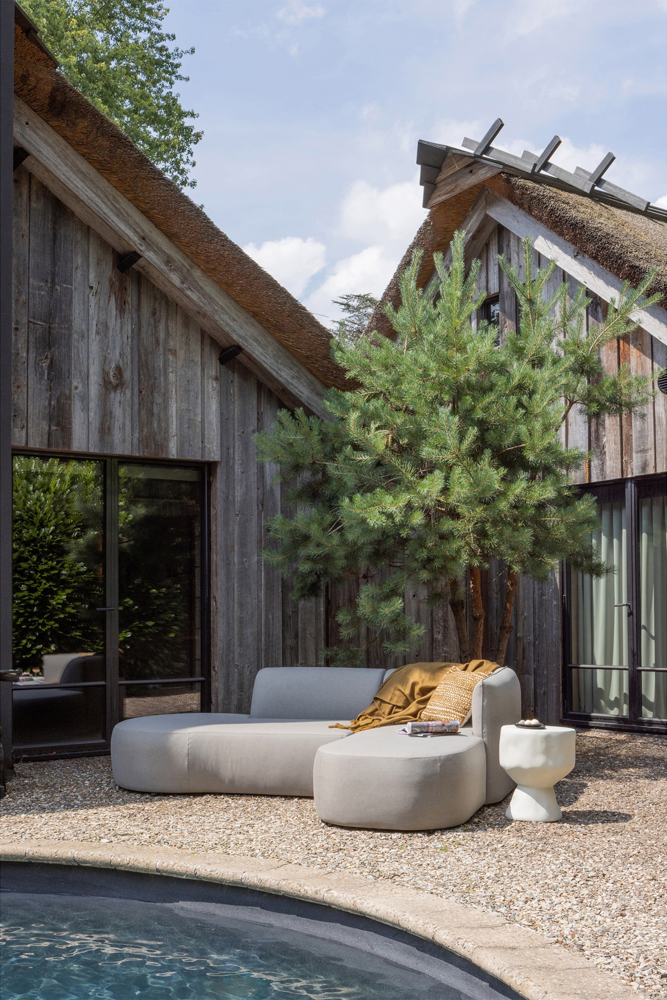 Grotere afbeelding bekijken: Een lichtgrijze gestoffeerde buitenloungebank toont de voorkant en rechterkant, met twee afgeronde, modulaire stukken: een banksectie en een loungesofa. Een mosterdgele plaid is over de bank gedrapeerd en een kleine witte sculpturale bijzettafel staat naast de chaise.
