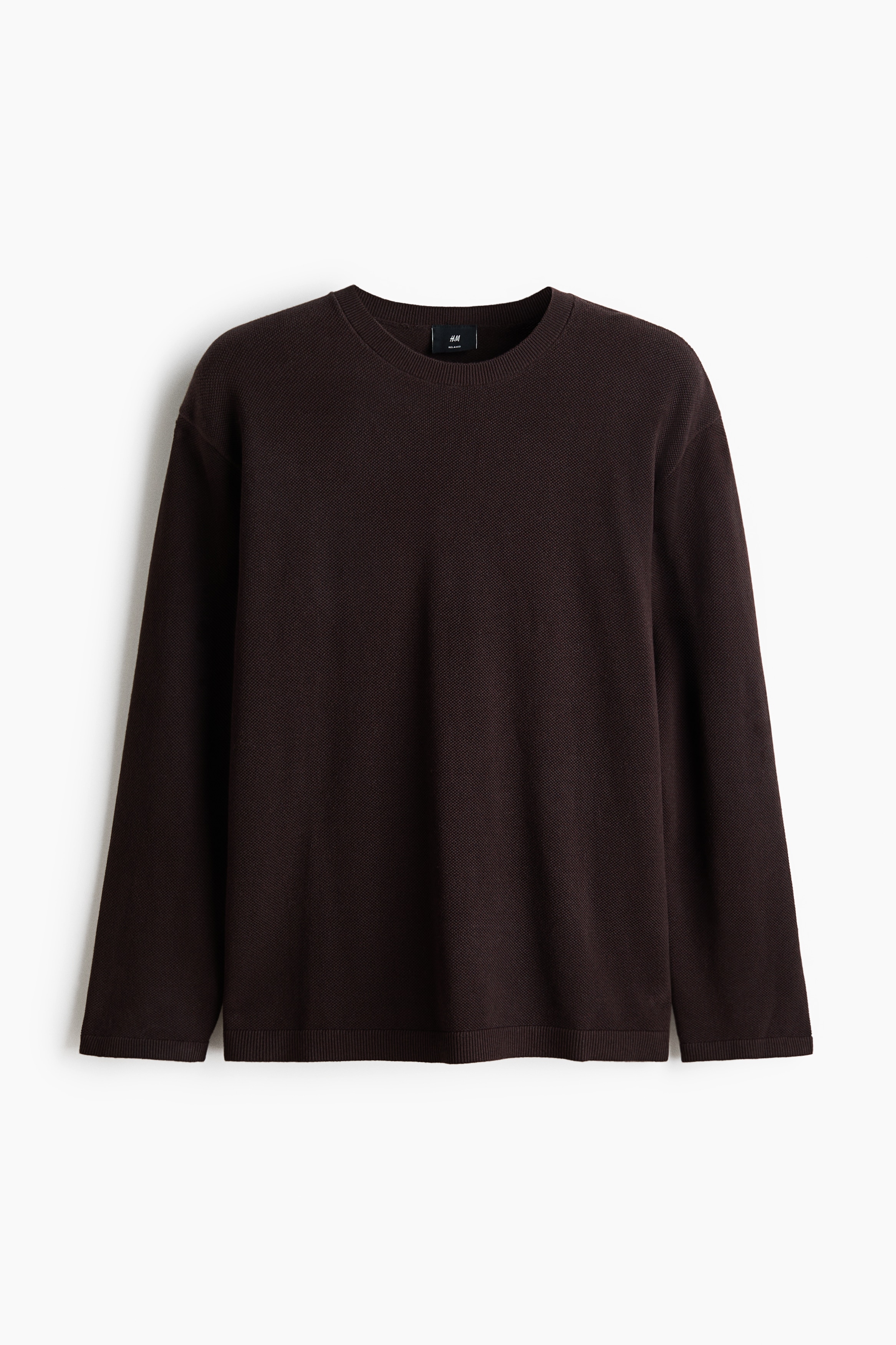 A dark brown textured knit sweater is being worn by the model, featuring a relaxed fit, a round neckline, and ribbing at the collar, cuffs, and hem. The sweater is paired with light brown trousers, with the model resting one hand in a pocket.