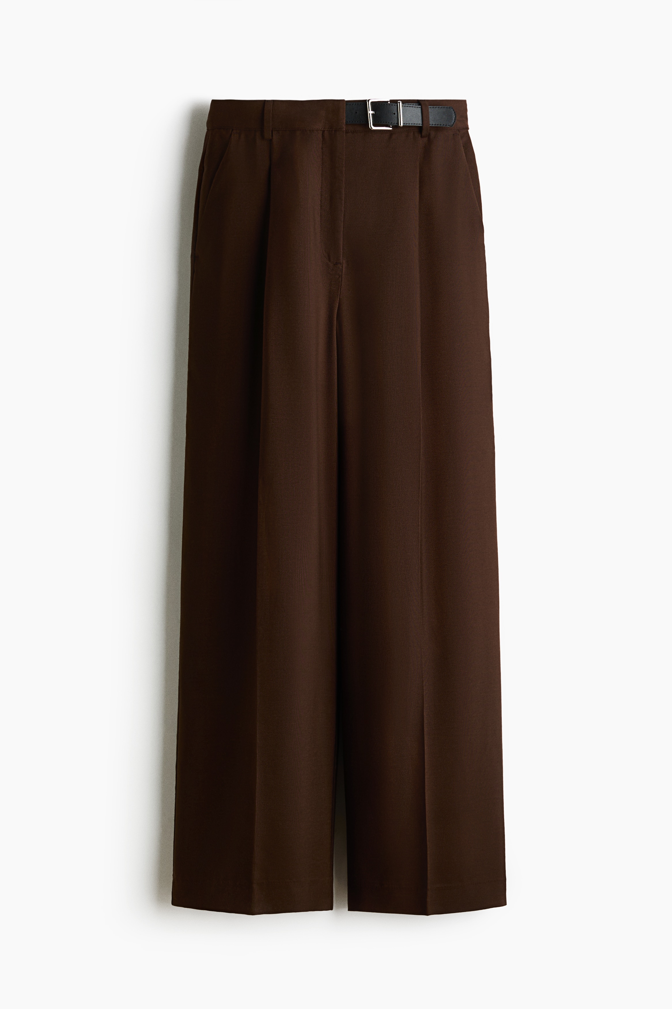 De model draagt een bruine wijde broek met persplooien aan de voorkant en staat met de handen in de zijzakken. De broek is gestyled met een ingestopte zwarte top met korte mouwen en opstaande hals en een zwarte riem met zilveren gesp.