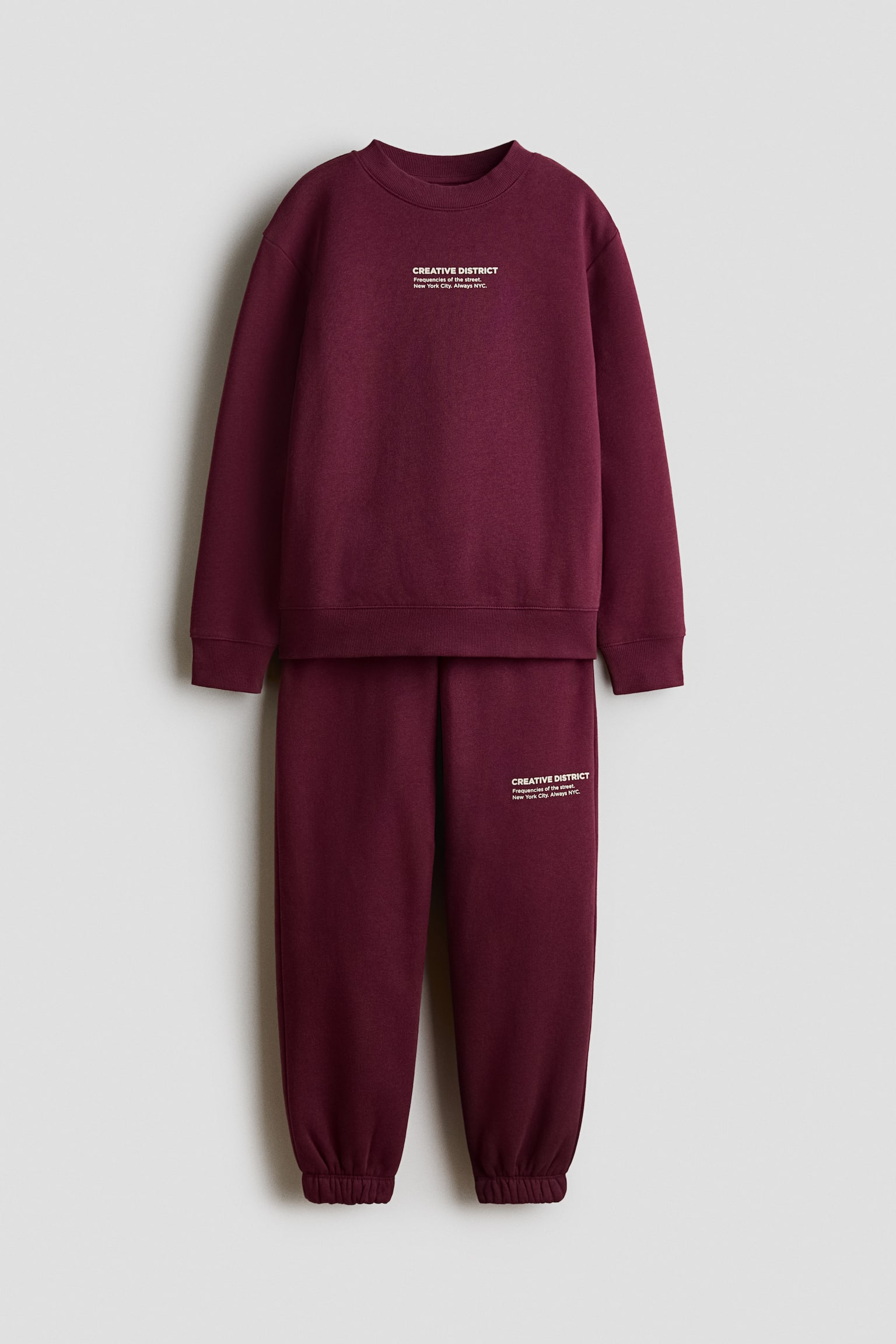 A burgundy two-piece set is shown from the front, featuring a crew-neck sweatshirt with ribbed cuffs and hem, and matching joggers with elasticated ankle cuffs. Both garments feature small white printed text on the chest and left thigh respectively.
