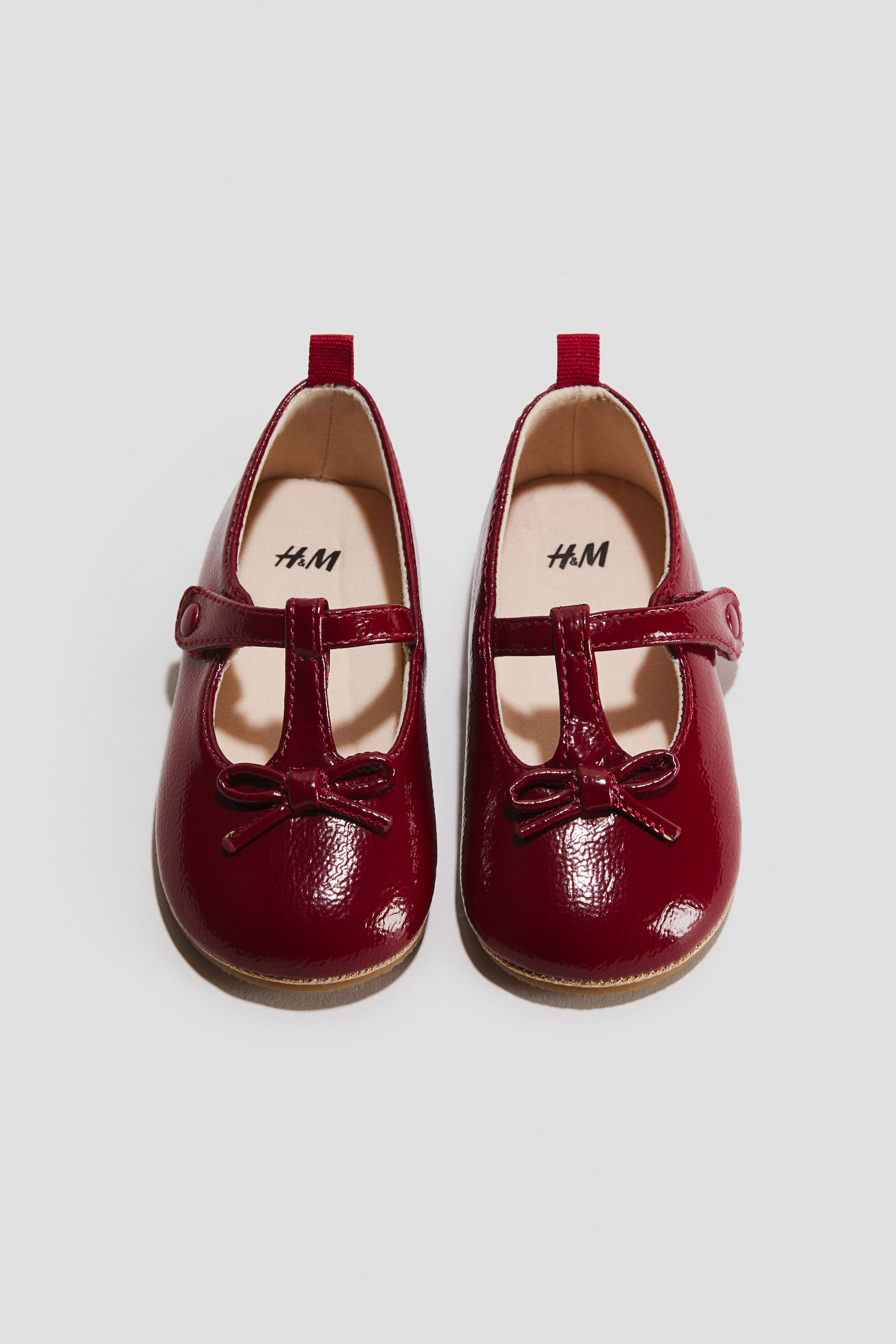 View larger image: A pair of deep red patent Mary Jane shoes is shown from a top-down angle, featuring rounded toes decorated with small bows and a T-strap design with hook-and-loop fastenings. The shoes display a glossy finish, light tan insoles, and small red pull tabs at the heels.