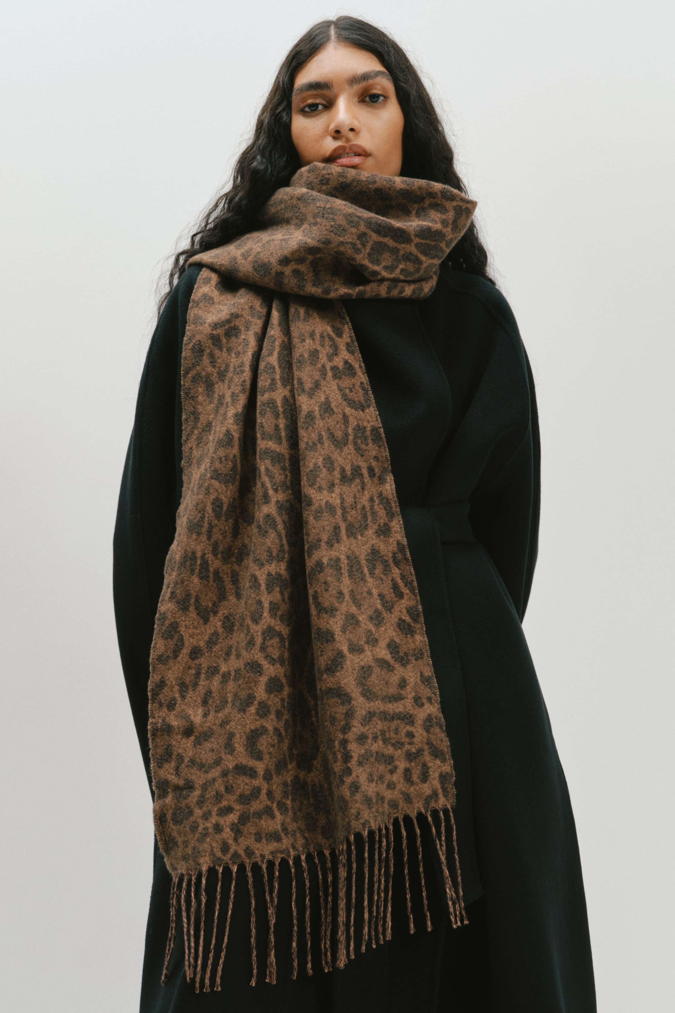 Ver imagen más grande: La modelo lleva una bufanda con estampado de leopardo, tejida en suaves tonos marrones y negros, con largas borlas en los lados cortos. La bufanda está enrollada alrededor del cuello y cae por la parte delantera sobre un abrigo negro.