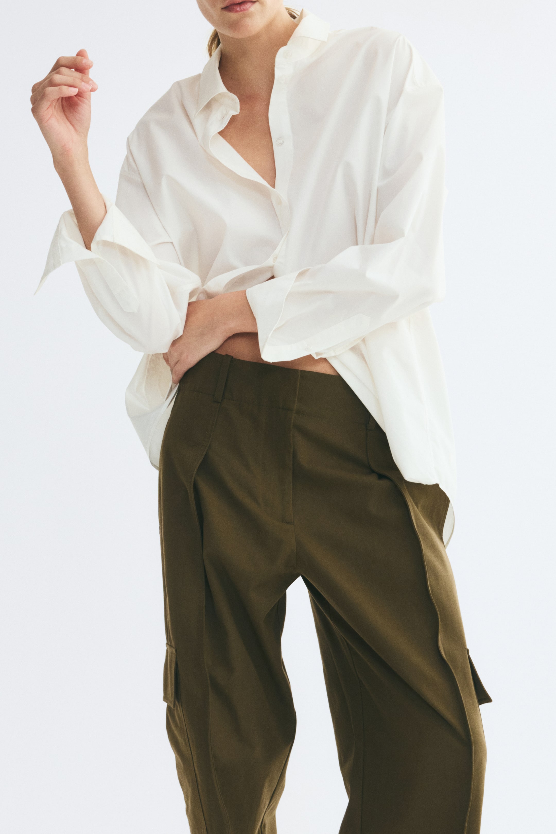 View larger image: Olive green cotton trousers are shown from the front in a high-waisted, barrel-leg fit. The trousers feature prominent front pleats, visible centre seams running down each leg, and side flap pockets, styled with a loose white shirt that is partly tucked in to reveal the waistband.