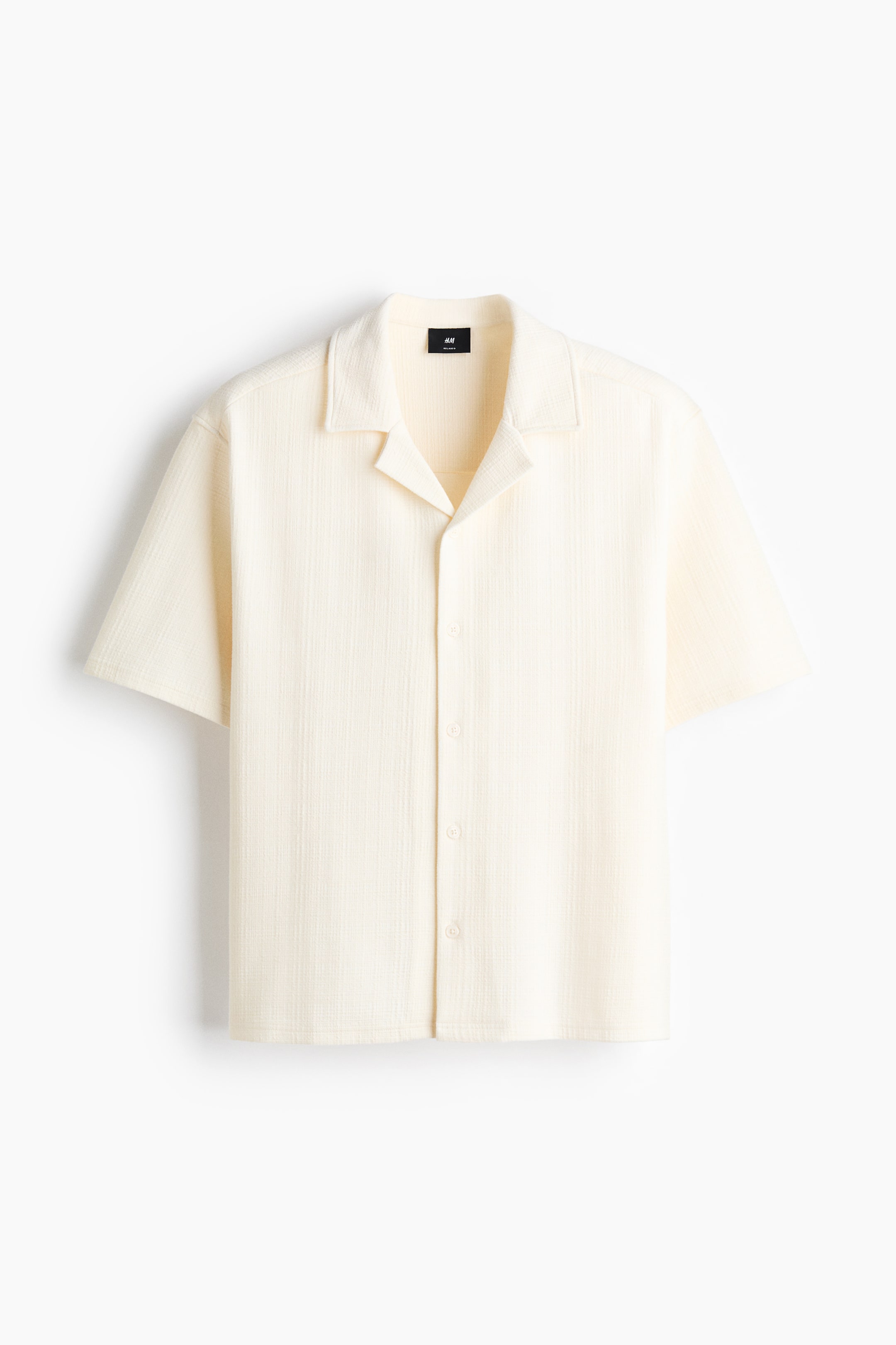 Visualizza immagine più grande: Una camicia a maniche corte color crema presenta un tessuto con una texture delicata. È caratterizzata da un colletto cubano e una chiusura frontale con bottoni tono su tono.