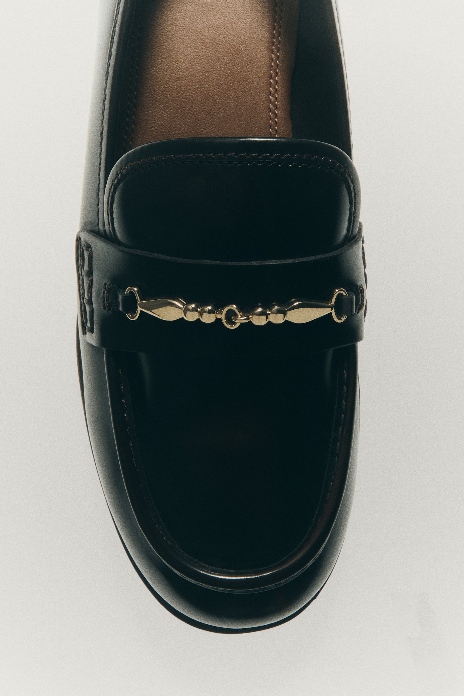 A pair of dark brown glossy loafers is showcased from a top-down perspective. Each shoe features a gold-toned chain detail across the vamp and a tan brown insole with visible branding. - 1