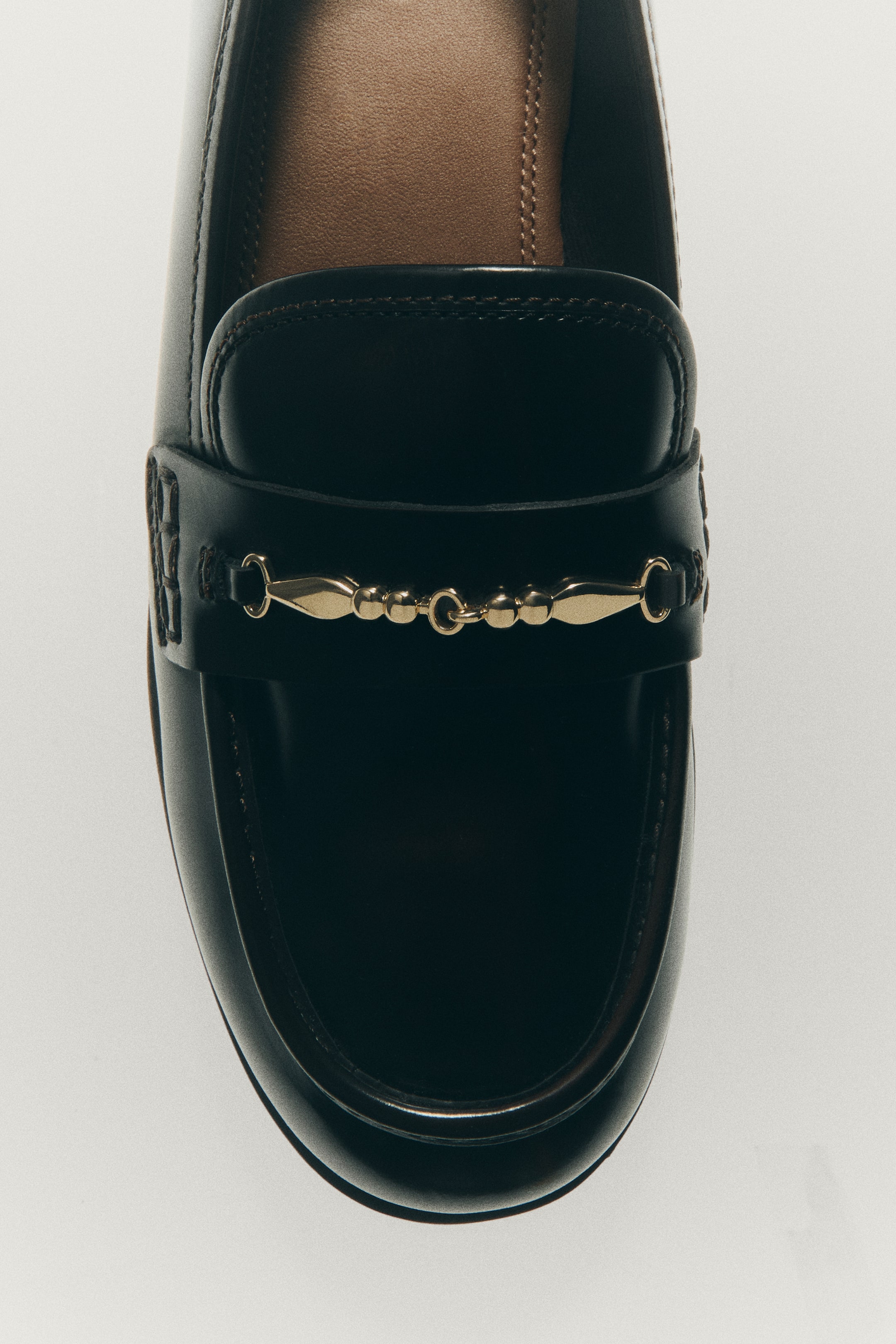 View larger image: A black leather loafer is shown from a top-down view, featuring a rounded toe with a moccasin seam and a leather strap accented with a gold-tone metal detail. The interior displays a contrasting brown leather lining and insole.