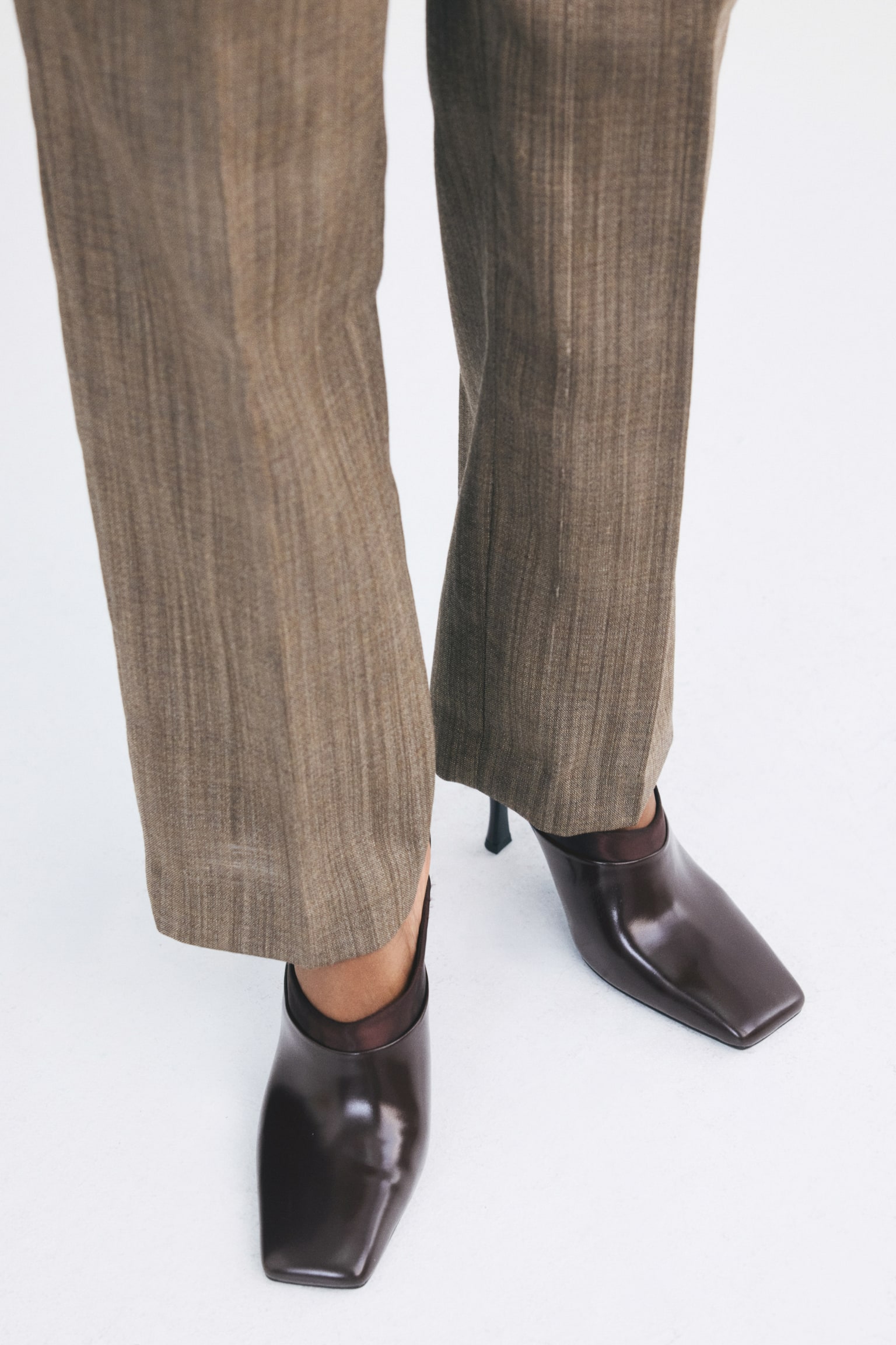 Mules in dark brown coated fabric and satin, featuring distinct square toes and stiletto heels, are being worn by the model. The shoes are styled under textured, light brown straight-leg trousers. - 1
