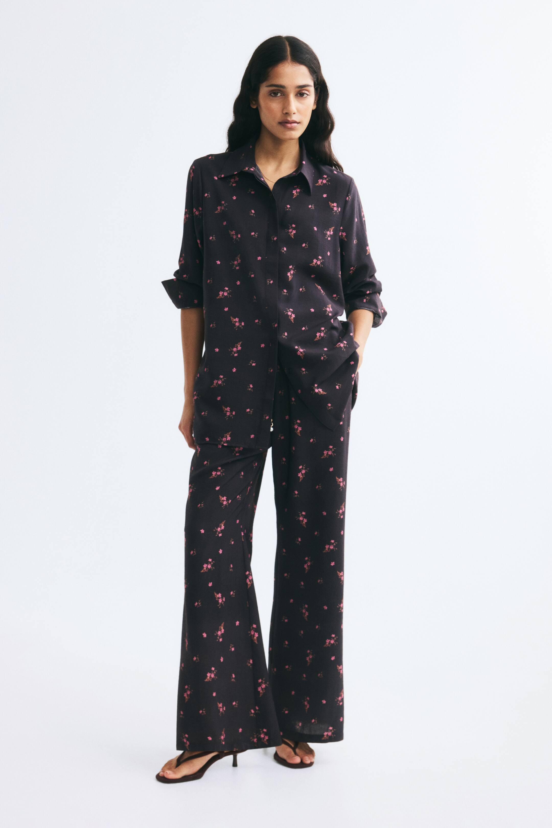 View larger image: A model is wearing the relaxed-fit, straight-leg pants from the front, showcasing their dark fabric adorned with a small pink floral print. The pants are styled with a matching long-sleeve shirt, with one hand resting in a diagonal side pocket to emphasise the fluid, relaxed drape.