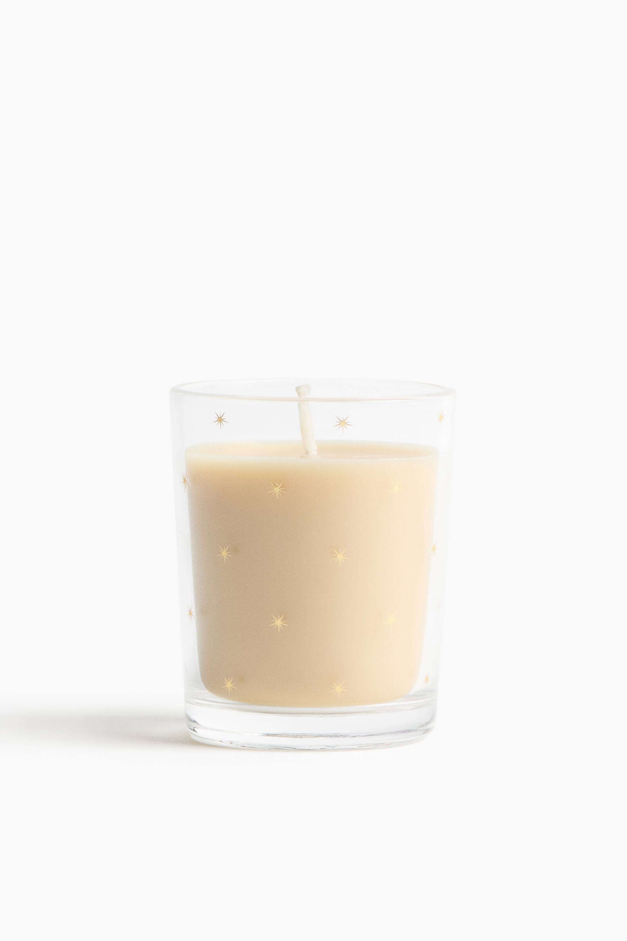 View larger image: A candle in a clear glass tumbler is standing upright, presenting a front view of its off-white wax and single white wick. The clear glass vessel features a pattern of small, scattered gold starbursts across its exterior.