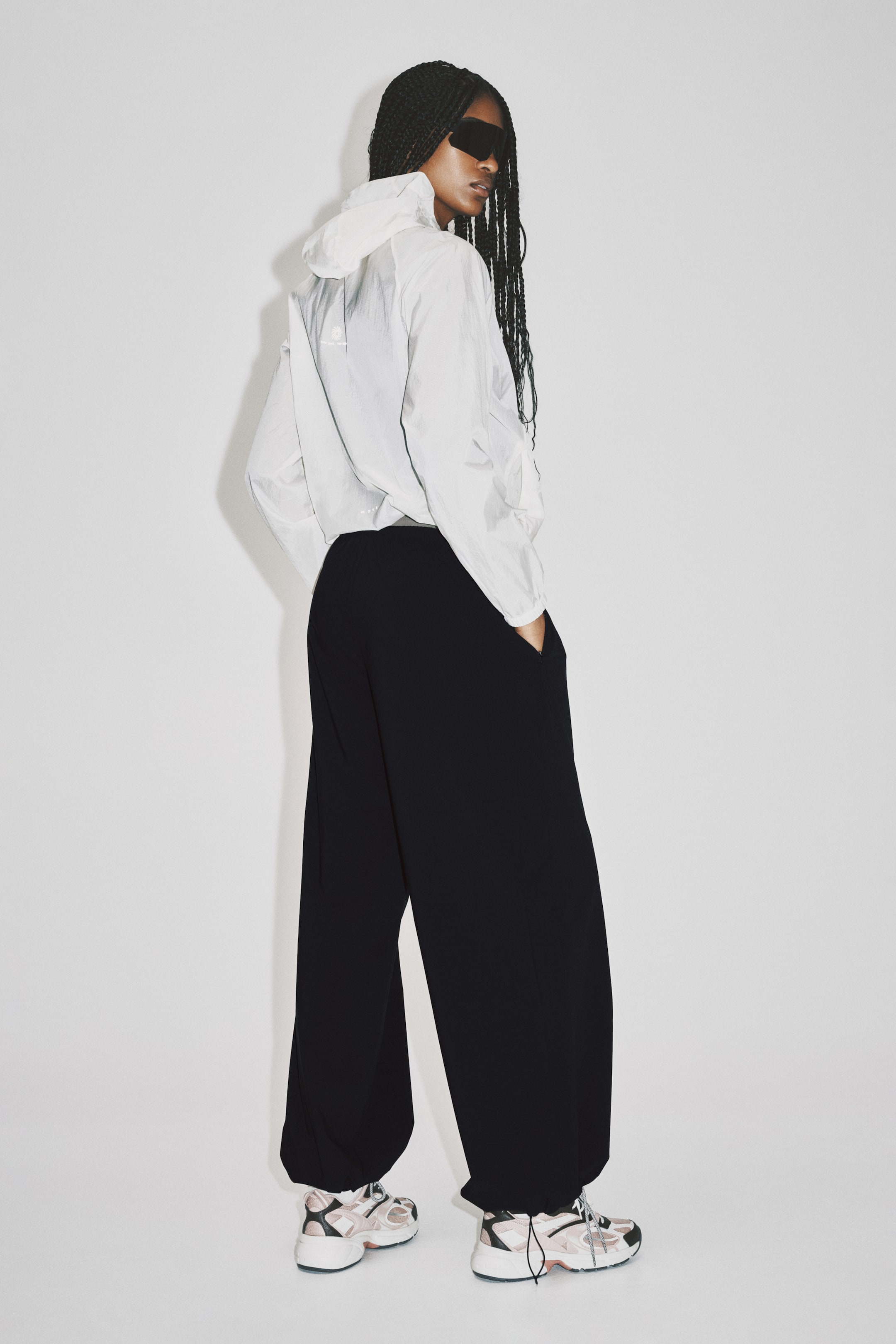 View larger image: Black wide-leg trousers feature a relaxed fit with cinched ankle hems, gathered above athletic shoes. The trousers showcase visible side pockets, with hands resting inside, and are presented from a rear-side angle.