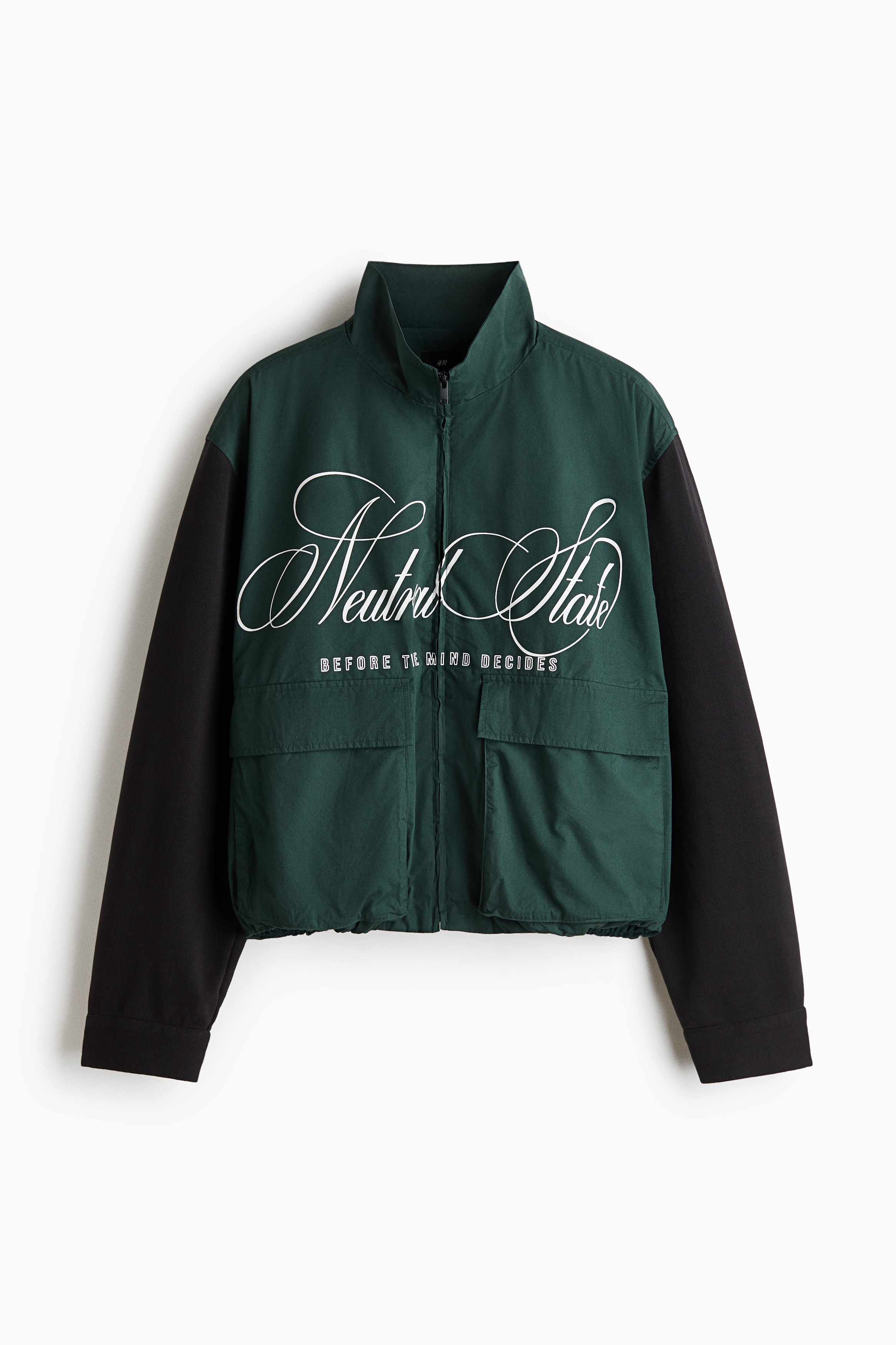 A colour-blocked jacket with a dark green body and black long sleeves is being worn by the model, featuring a stand collar, a front zipper, and large front patch pockets with flaps. The jacket has a white script print reading "Neutral State" across the chest and is layered over a white T-shirt, styled with black pants and a black cap.