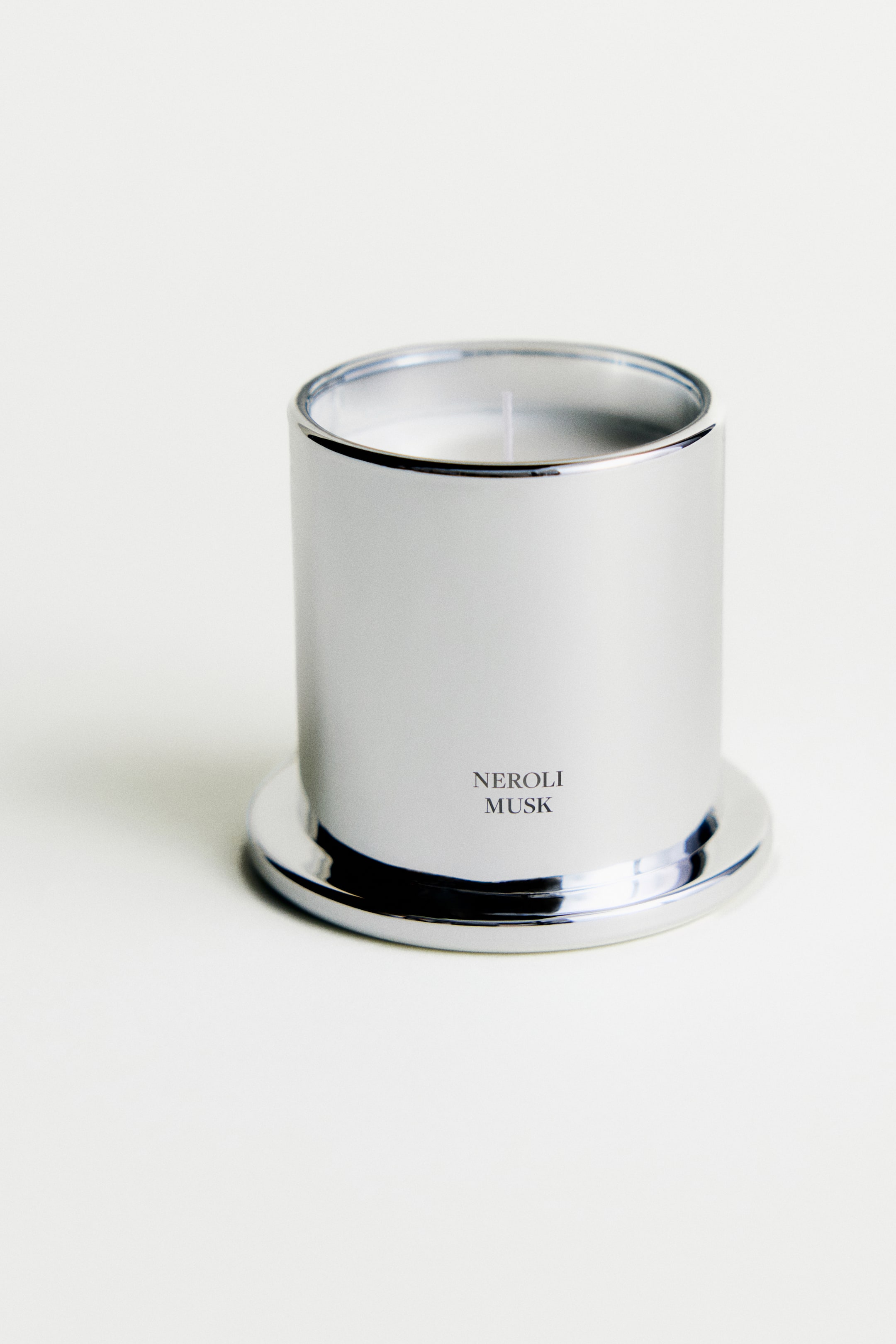Se større billede: Et frontalt billede, der viser et duftlys i en cylindrisk glasbeholder med en reflekterende sølvfarvet kromfinish, hvor teksten "NEROLI MUSK" er trykt med sort skrift på forsiden. Lyset hviler på sit matchende kromklokkelåg, som fungerer som en base, og afslører hvid voks med en enkelt væge.