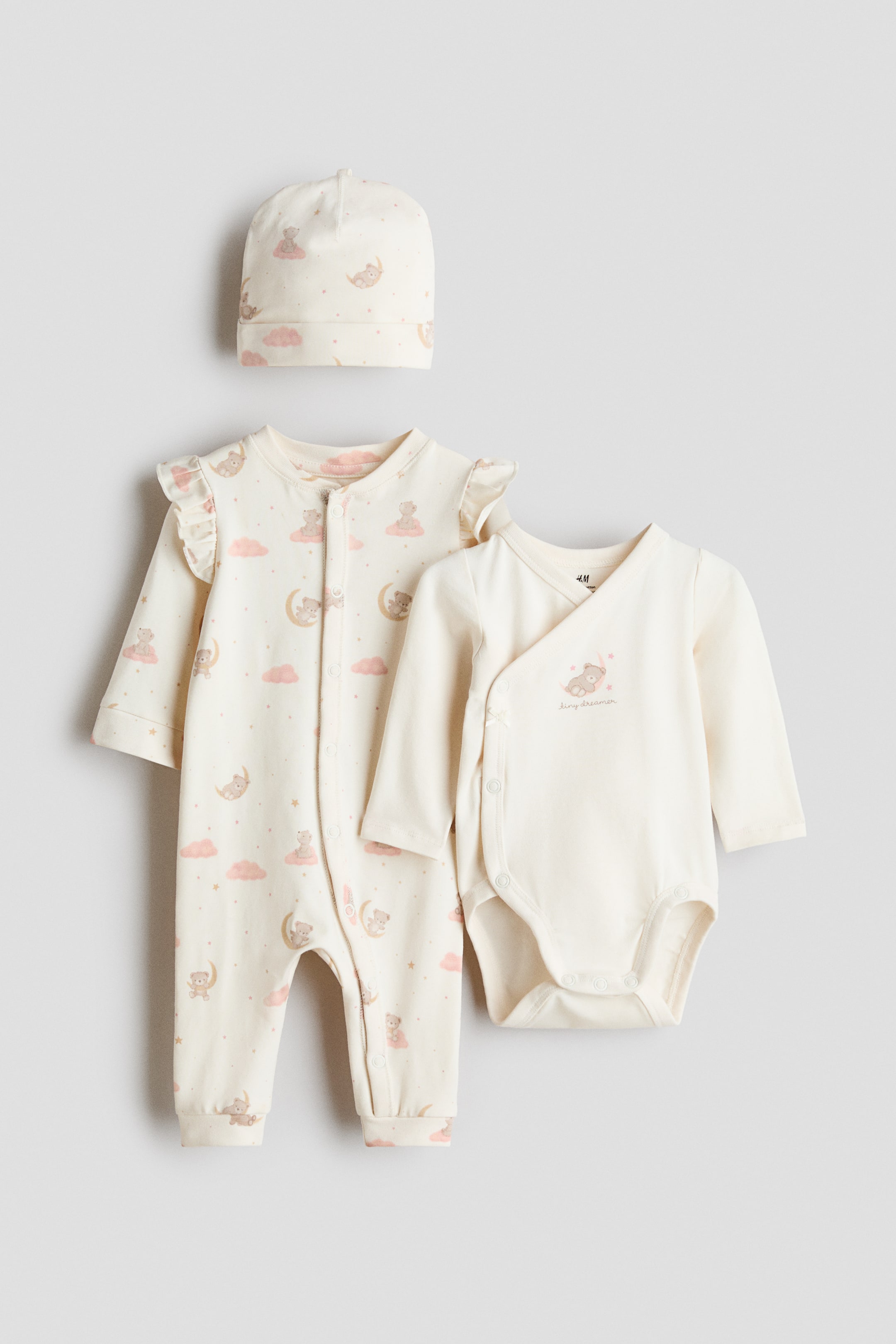 View larger image: A three-piece baby set is showcasing a front view, comprising a long-sleeved sleepsuit, a long-sleeved bodysuit, and a beanie. The sleepsuit and beanie are in a cream hue adorned with a pattern of sleeping bears on crescents and pink clouds, while the bodysuit is plain cream featuring a small sleeping bear graphic on the chest. The sleepsuit is crafted with a trimmed neckline, ruffled shoulders, press-studs down the front and along one leg, and wide trims at the cuffs and hems; the bodysuit displays a wrapover front with press-studs at the sides and bottom; and the beanie exhibits a turn-up at the hem.